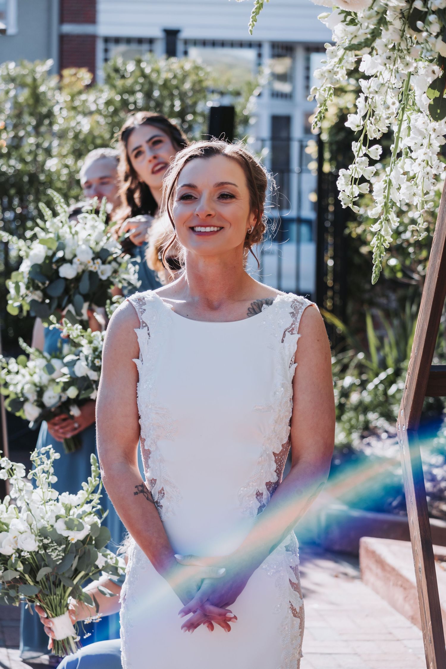 Radiant bride in white lace gown smiles at altar during outdoor wedding ceremony with rainbow lens flare effect.