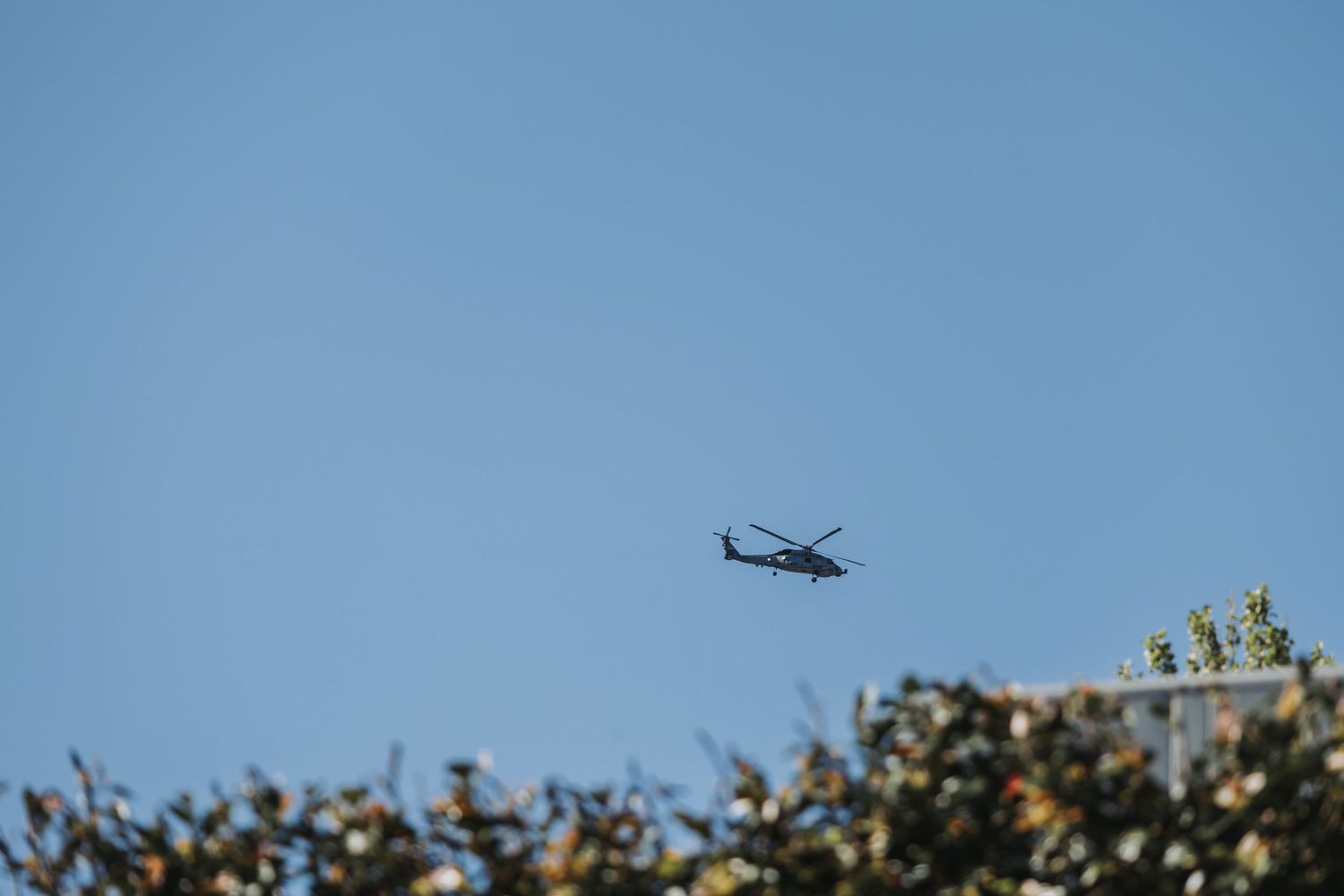 Black helicopter flying over treetops against clear blue sky.