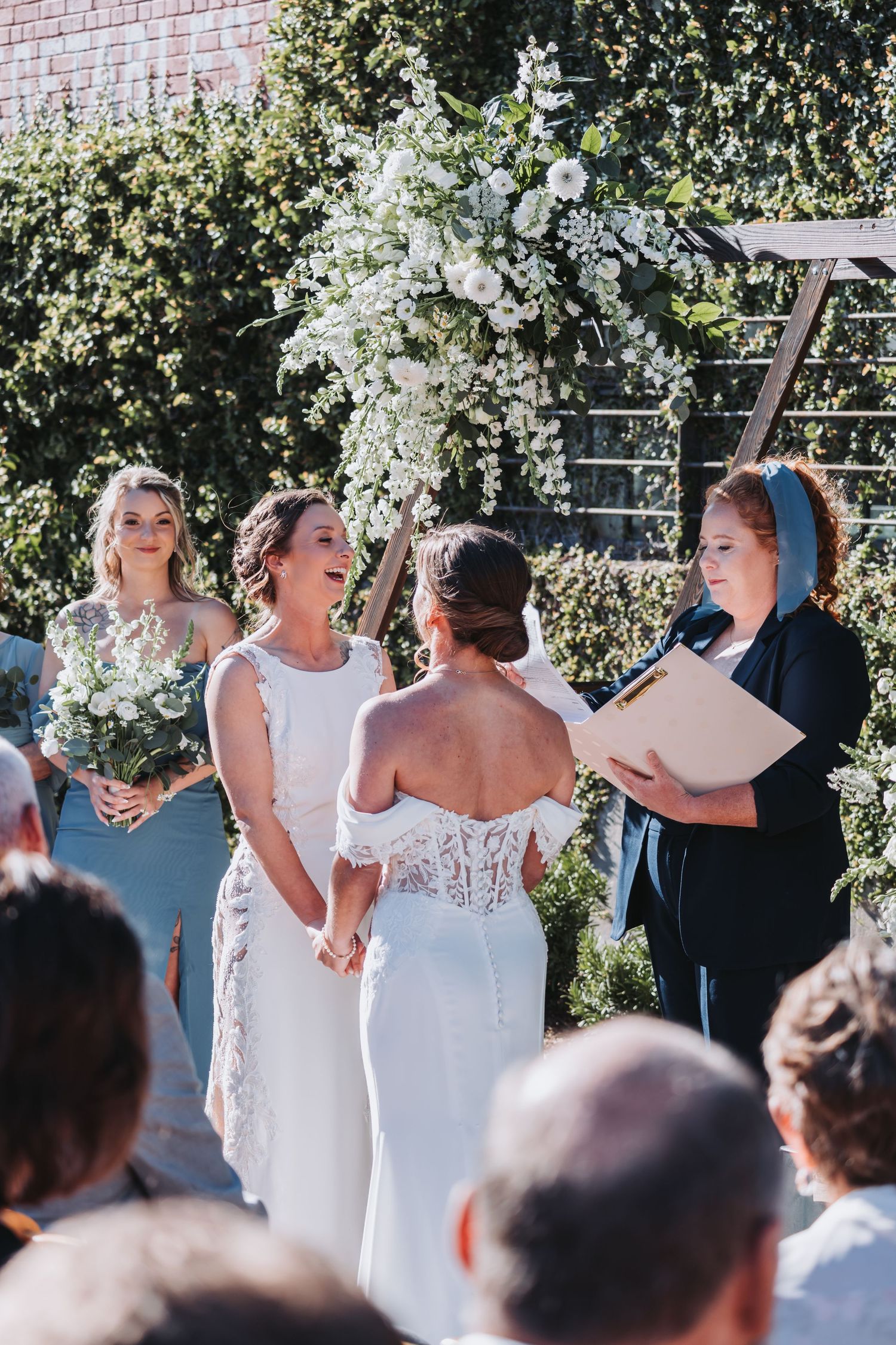 Two brides hold hands at outdoor wedding ceremony with white floral arch and bridesmaid in blue dress.