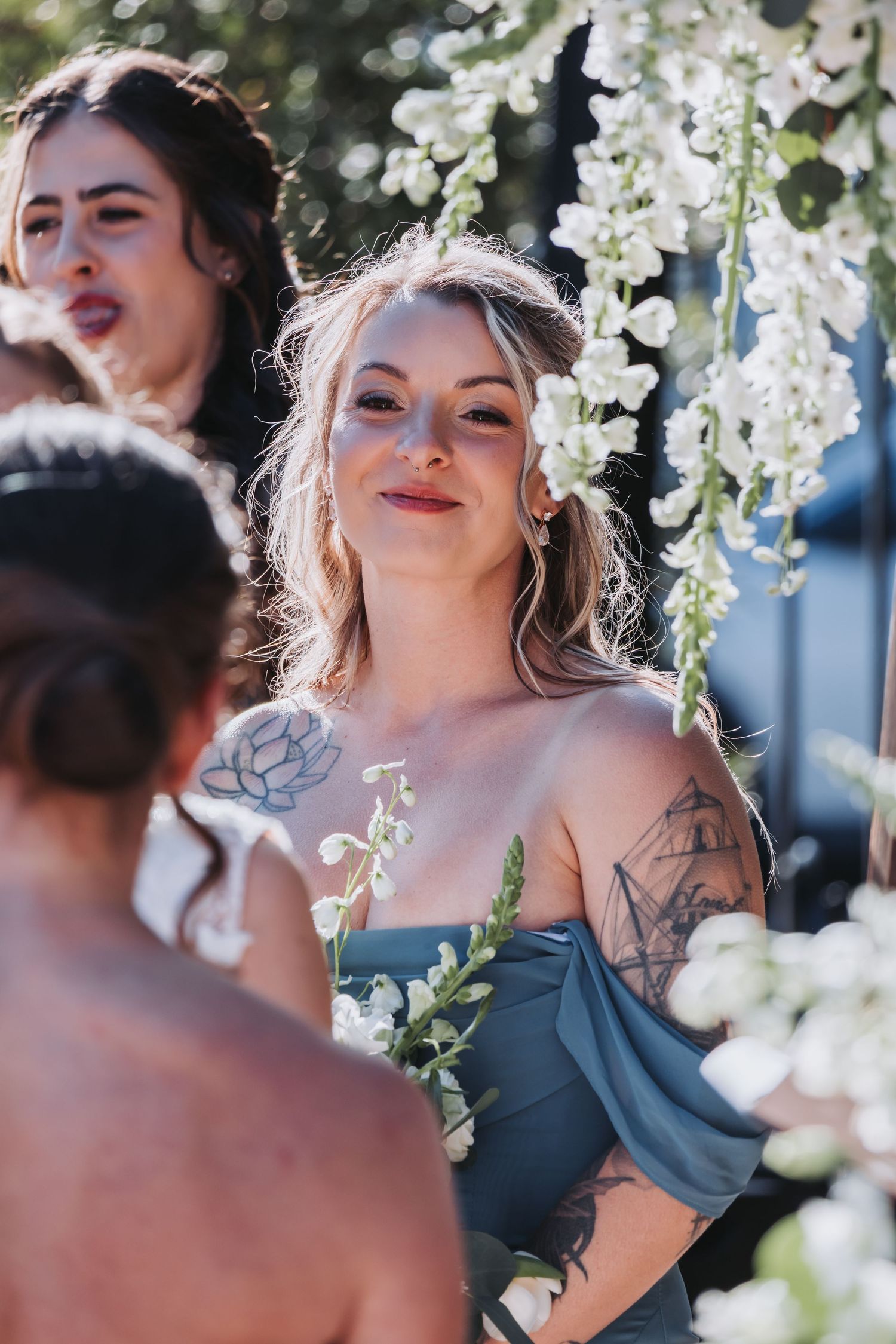 Tattooed bridesmaid in blue off-shoulder dress holds white flowers at a sunlit outdoor wedding ceremony.