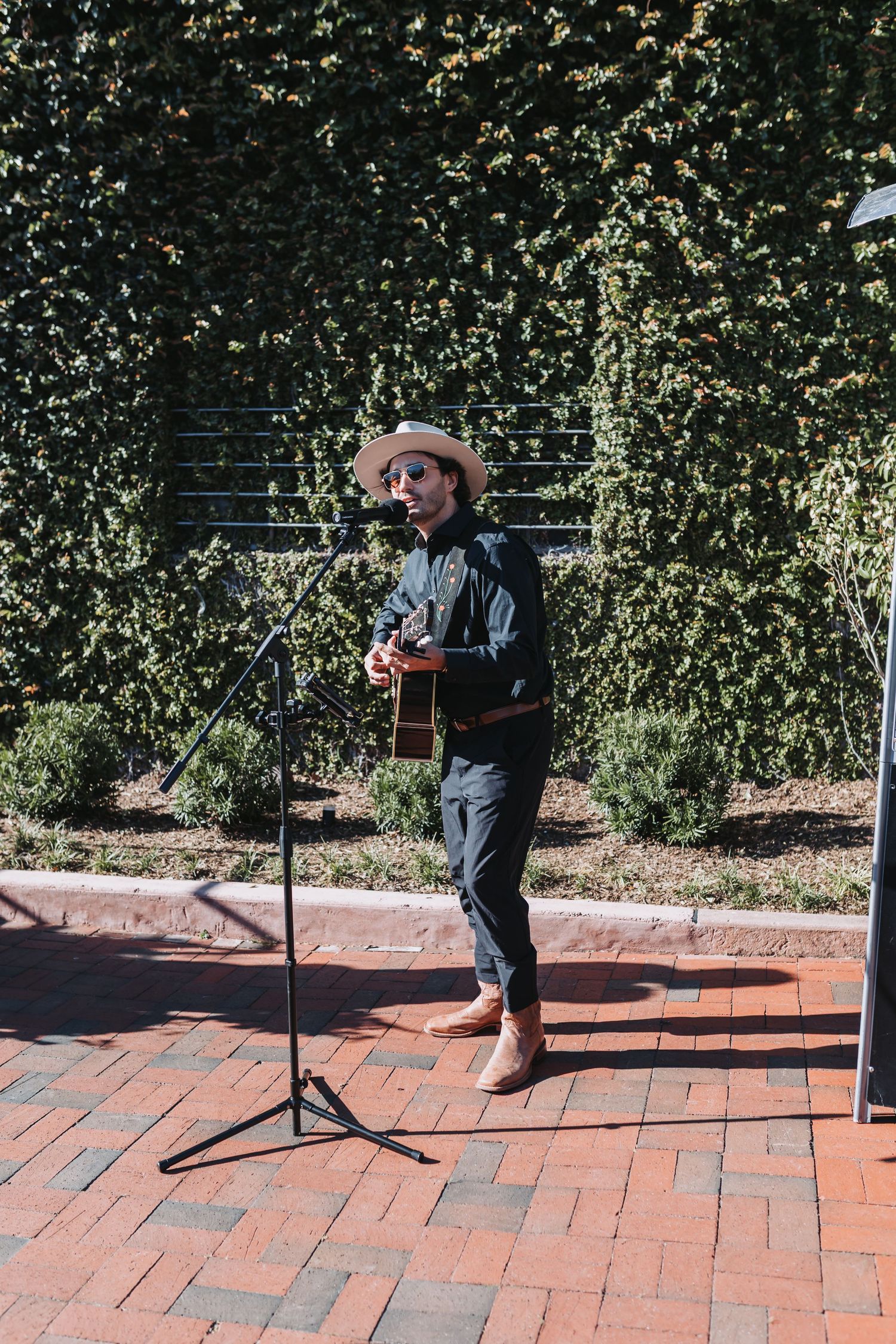 Musician in straw hat plays acoustic guitar at outdoor wedding ceremony on brick patio.