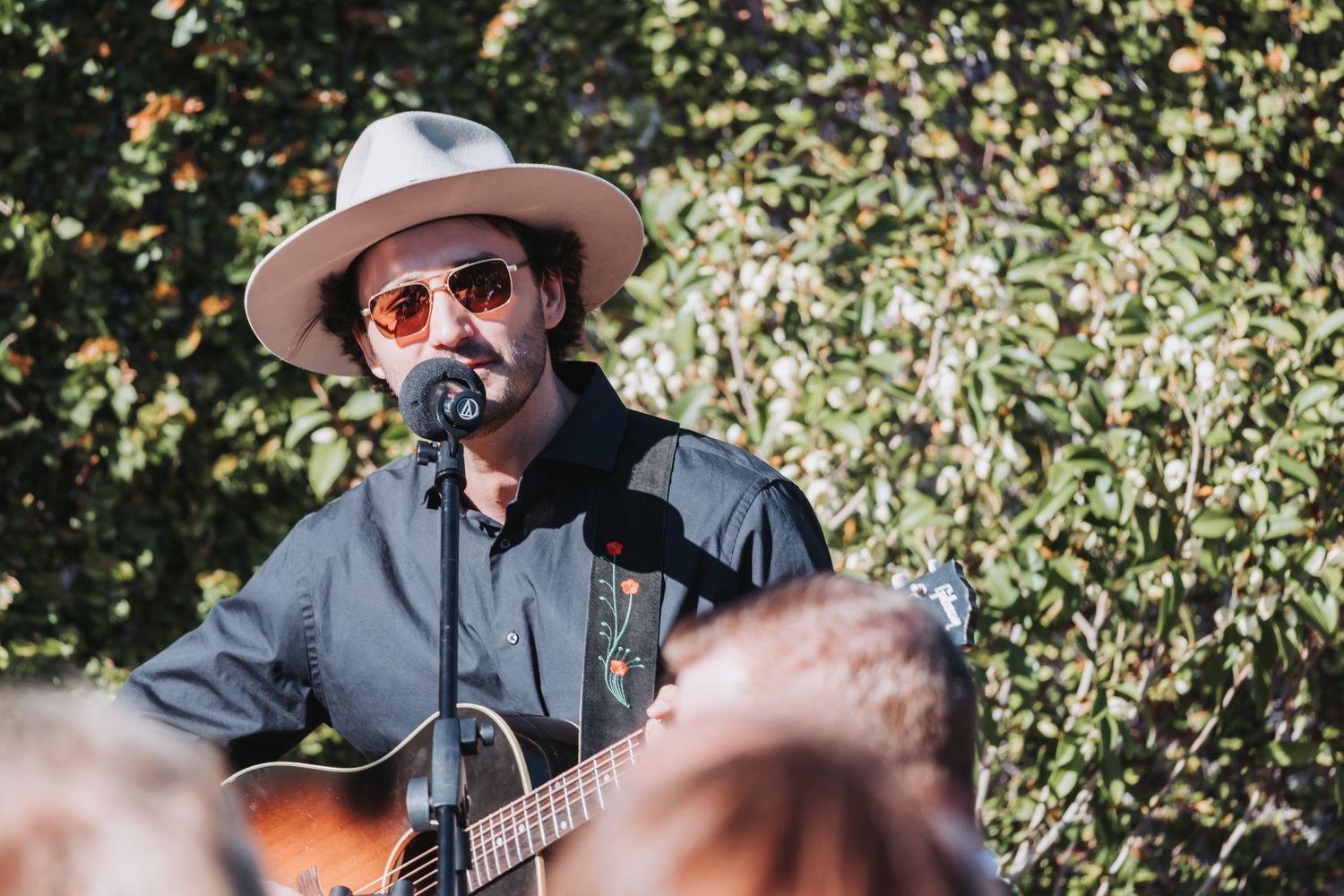 Musician in a wide-brim hat and sunglasses plays acoustic guitar at an outdoor wedding reception.