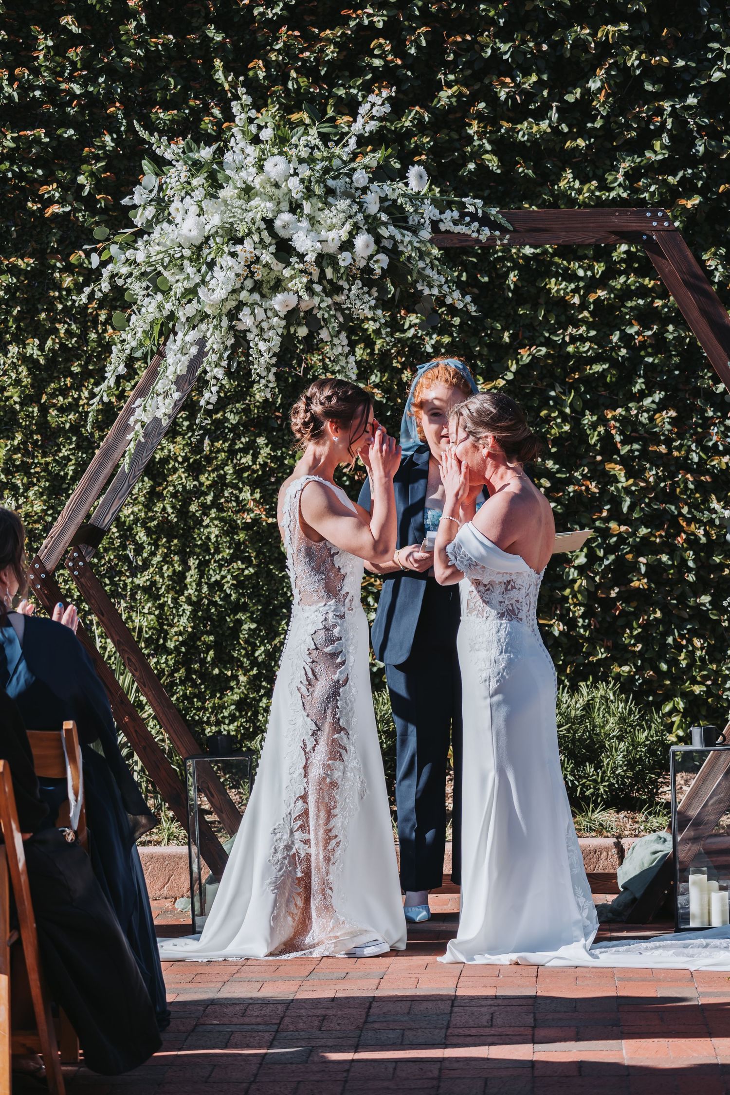 Two brides exchange vows under a floral arch at an outdoor wedding ceremony.