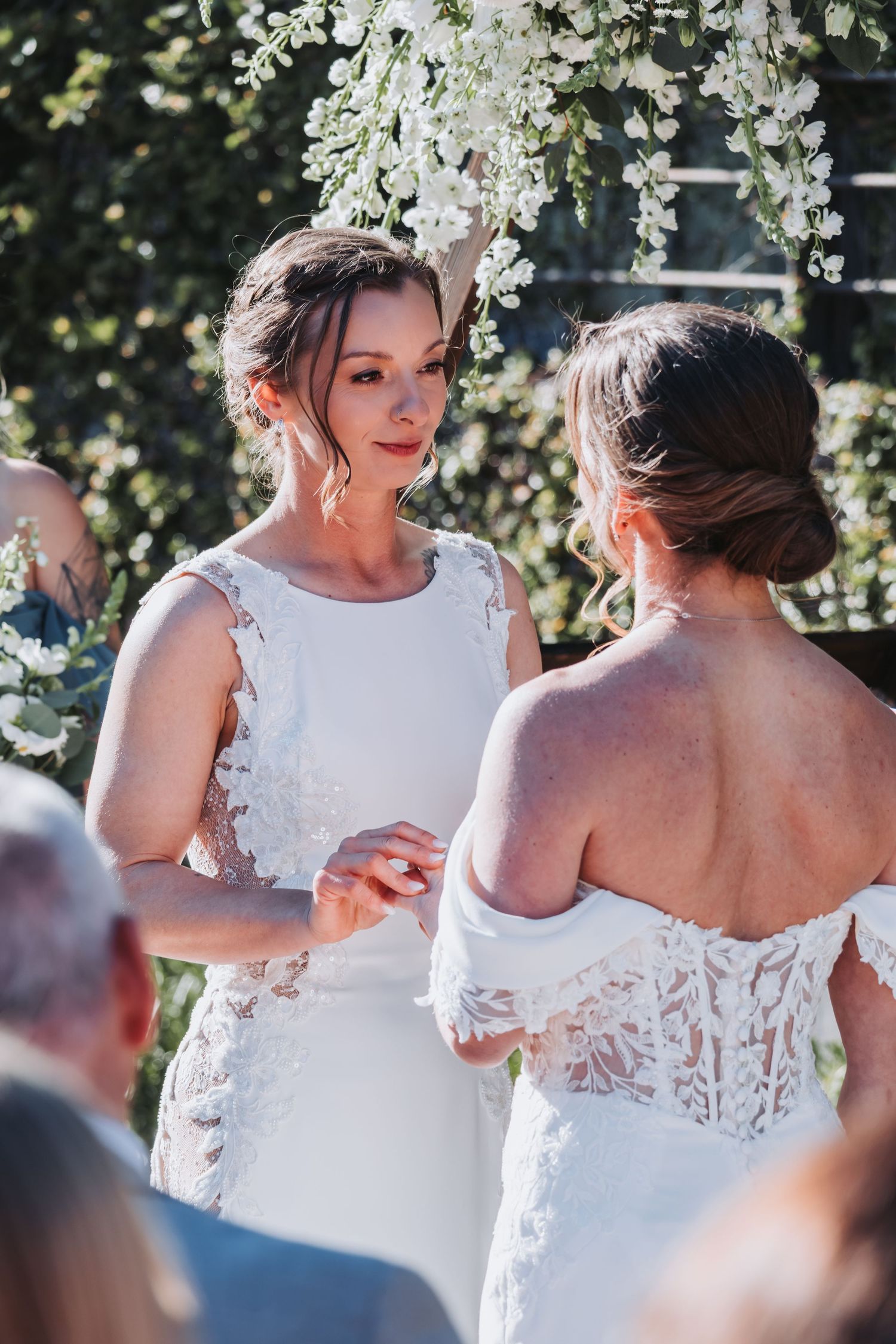 Two brides holding hands during intimate ring exchange moment at outdoor wedding ceremony.