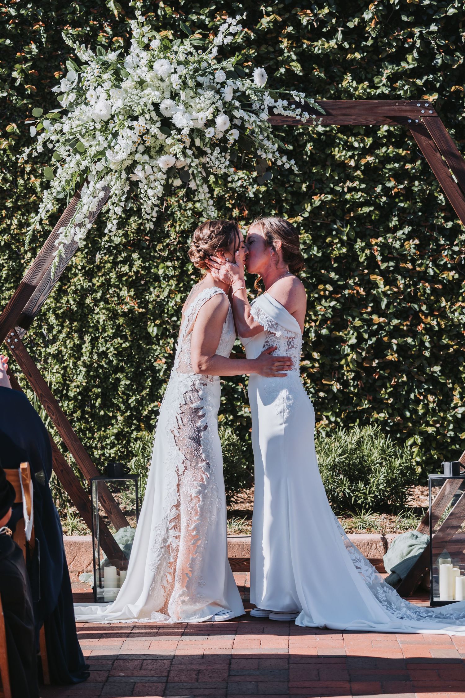Two brides in white gowns share a kiss under a wooden arch adorned with white flowers at an outdoor wedding ceremony.