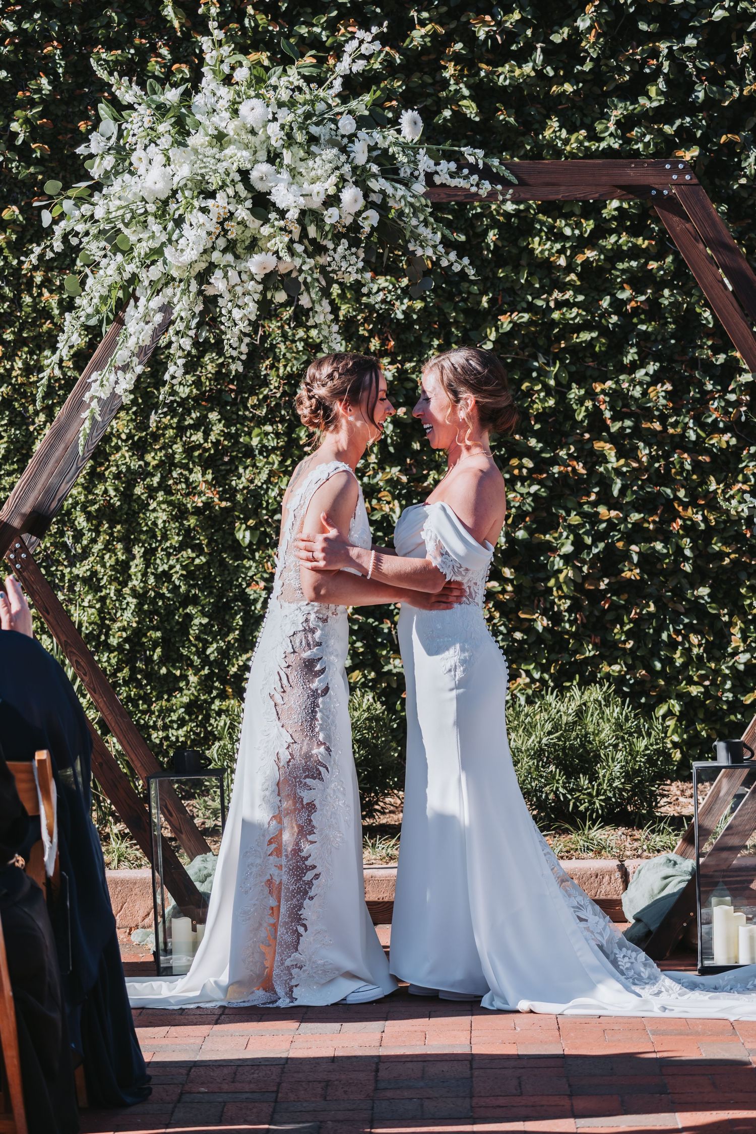 Two brides in white gowns embrace under a floral arch at an outdoor wedding ceremony.
