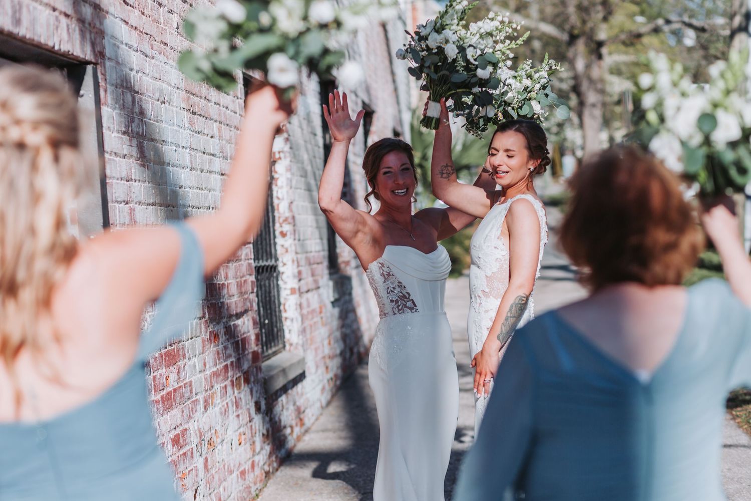 Two brides in white gowns celebrate with bridesmaids in blue dresses, holding bouquets near a brick wall.