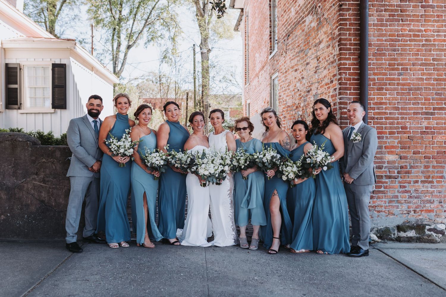 Wedding party in dusty blue dresses and gray suits posing together outdoors near a brick building.