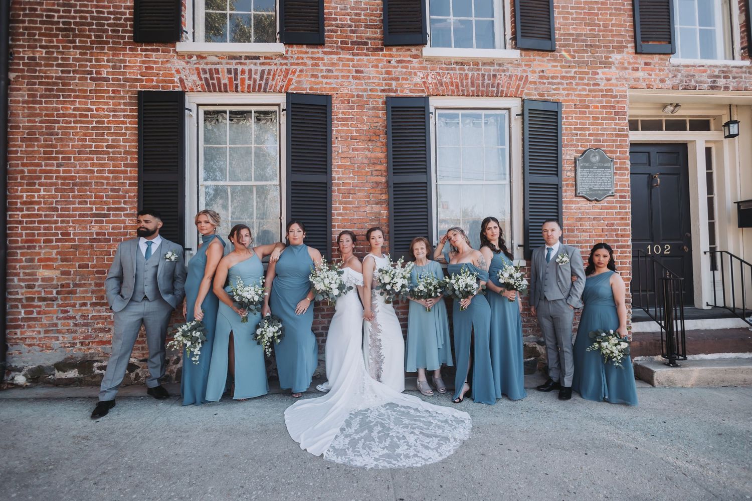 Wedding party poses in front of brick building, bridesmaids in blue gowns and groomsmen in gray suits flanking the bride and groom.