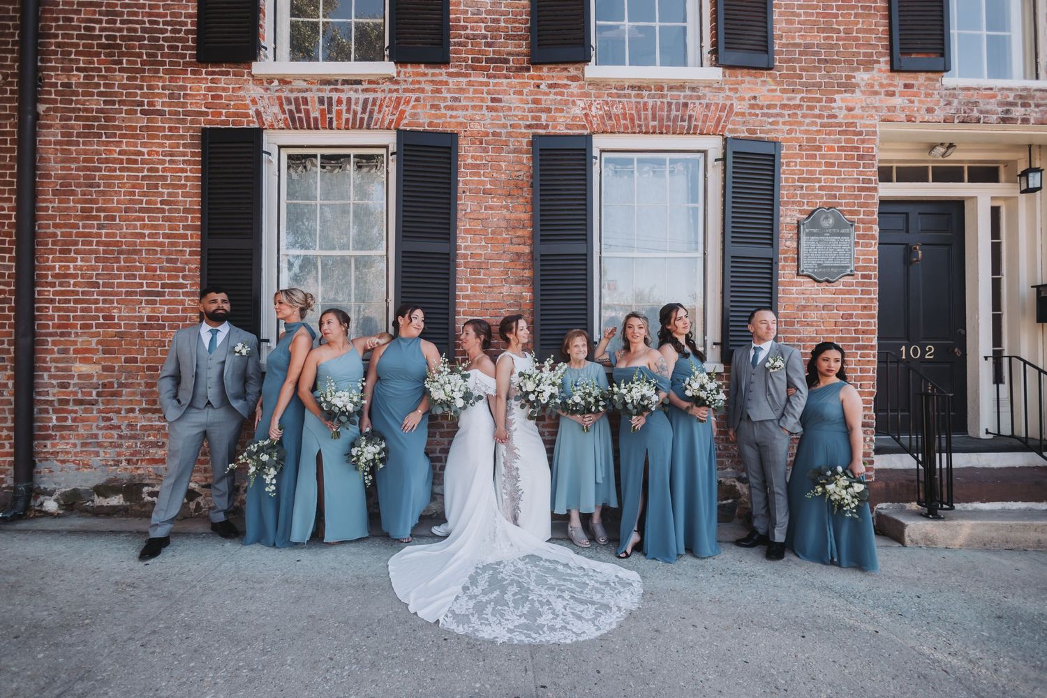Wedding party in dusty blue gowns and gray suits posing before a brick building with black shutters.