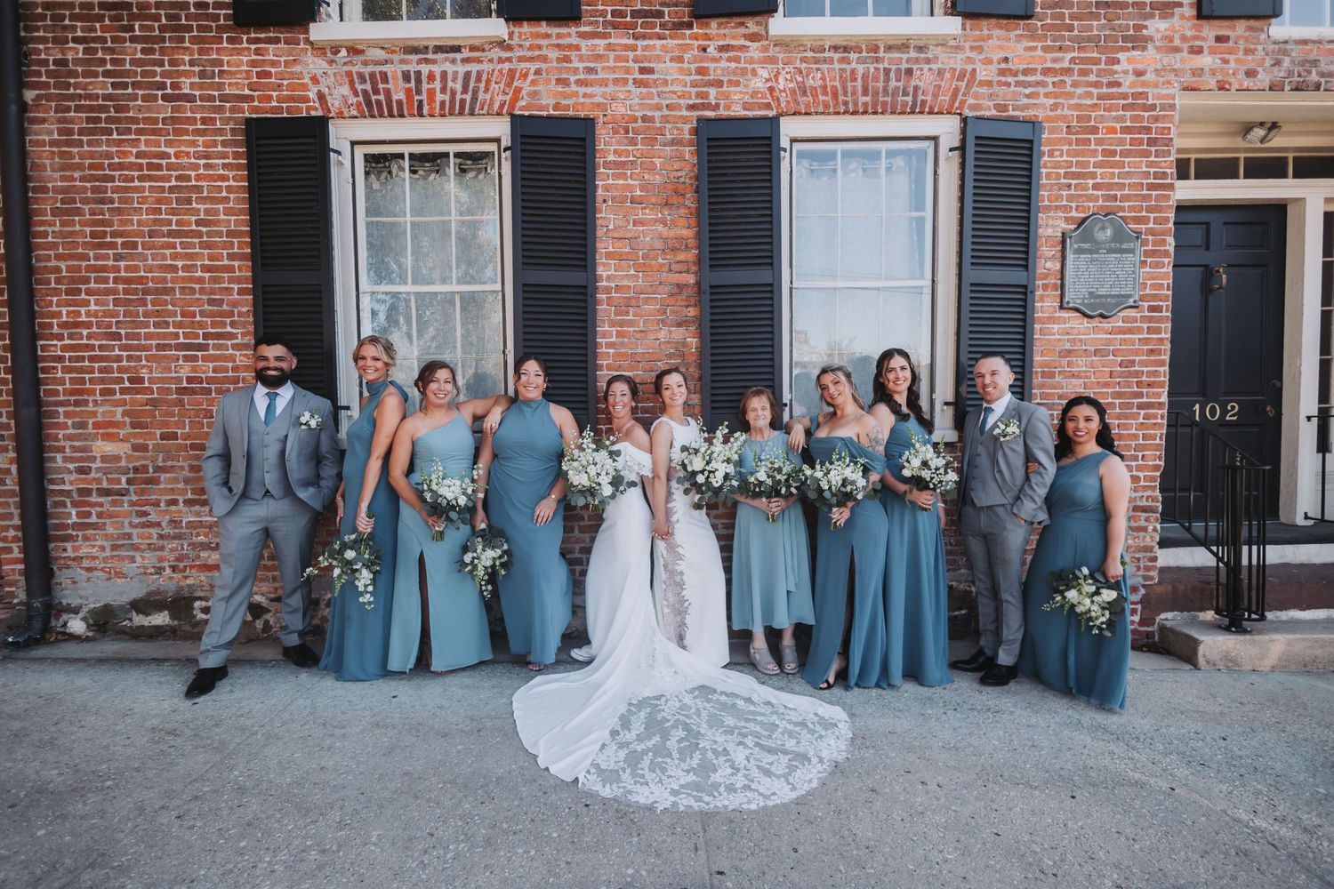 Wedding party poses in dusty blue dresses and gray suits before brick building with black shutters.