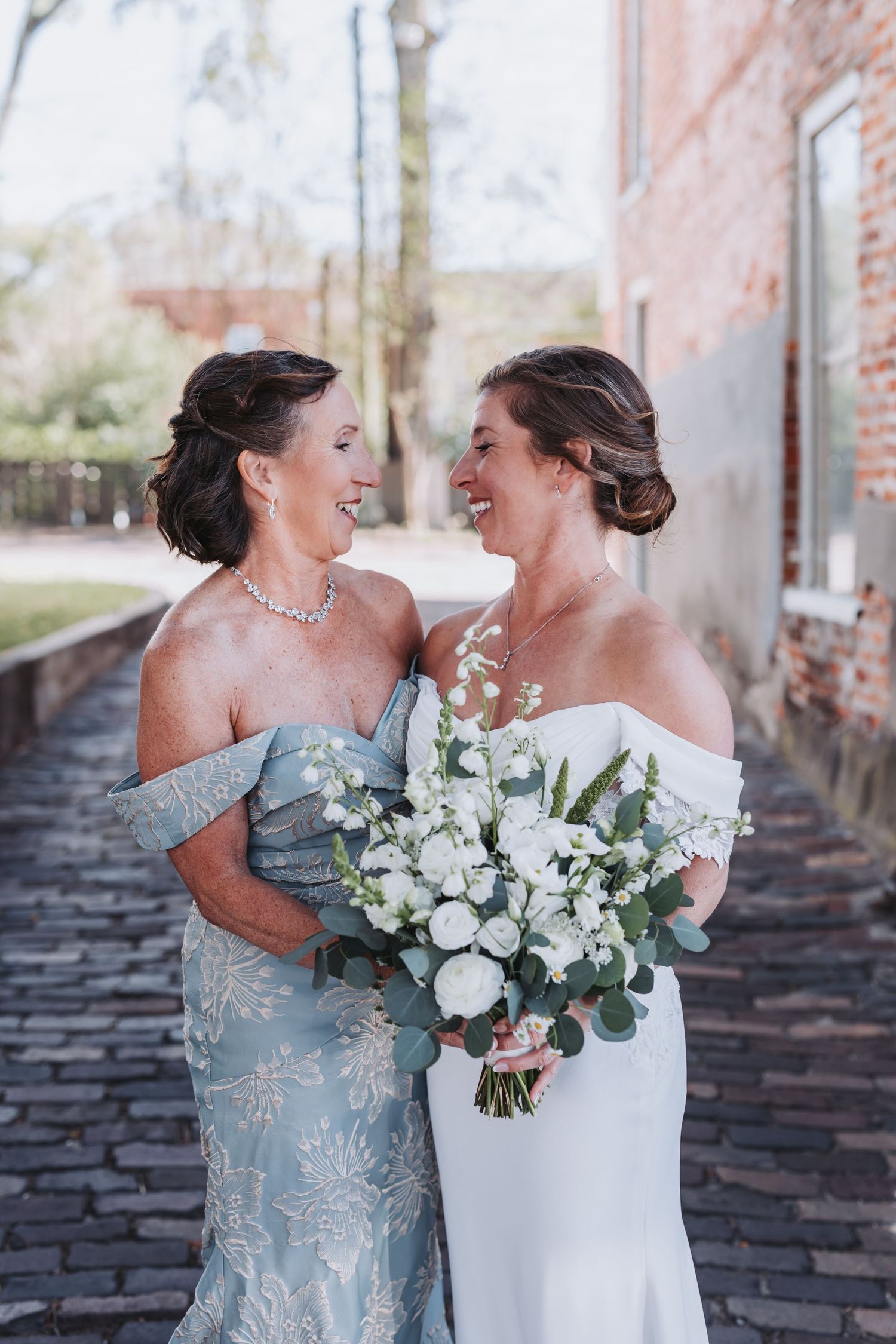Bride in white off-shoulder gown and mother in blue lace dress smiling at each other, holding white floral bouquet outdoors.