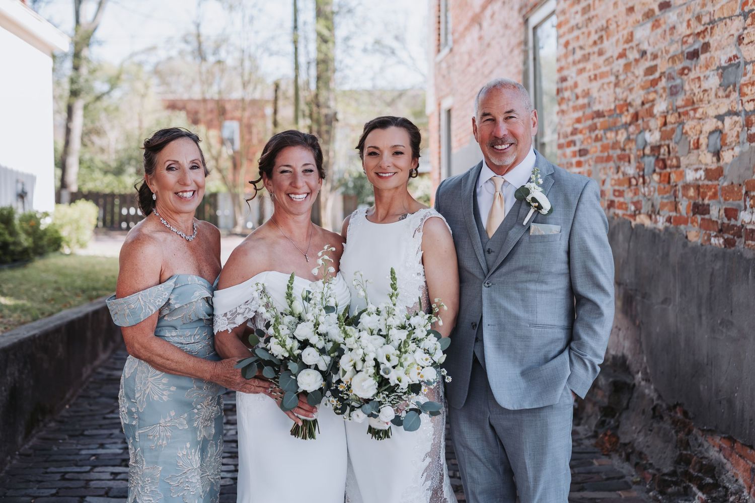 Bride posing with family members in blue-gray attire, holding white floral bouquet outside brick building.