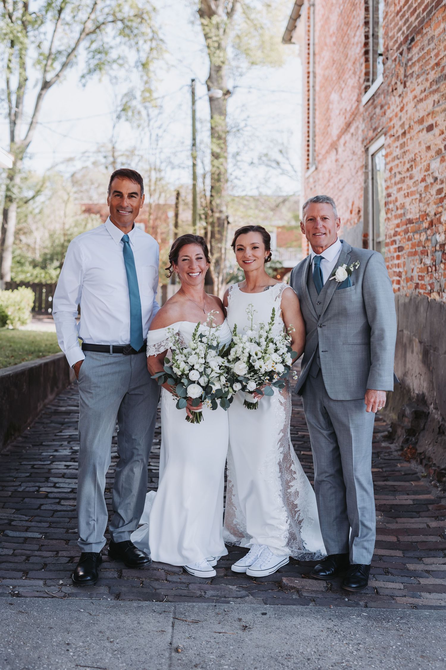 Two brides in white gowns holding bouquets pose with two men in suits on a cobblestone path by a brick building.