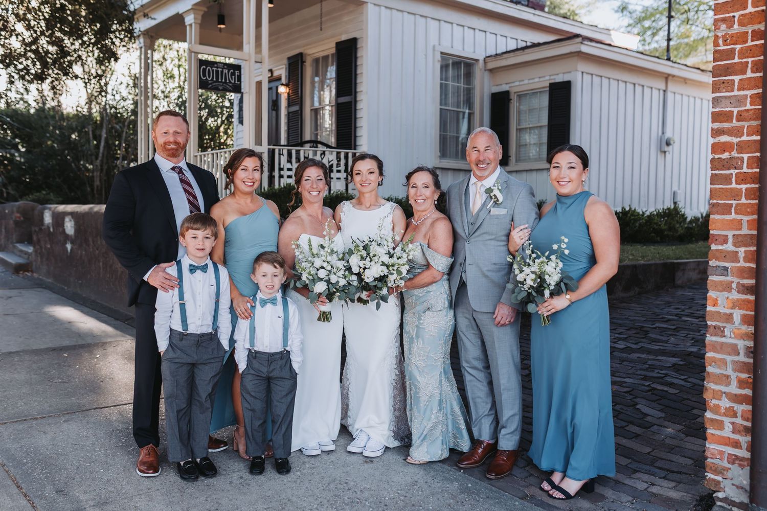 Wedding party in blue attire poses together outside a white building on a sunny day.
