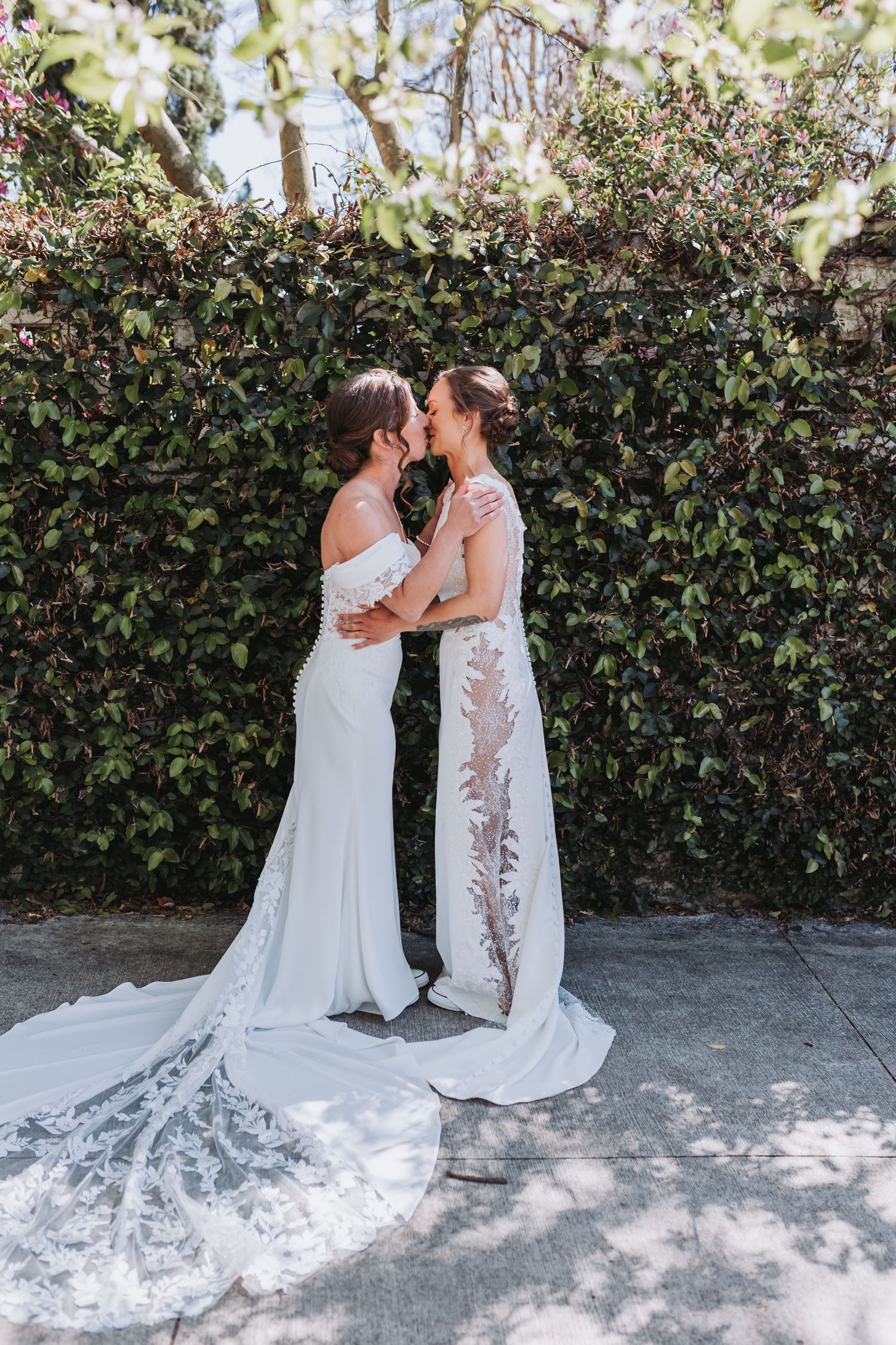 Two brides in white lace wedding gowns share a kiss in front of a lush green hedge wall.
