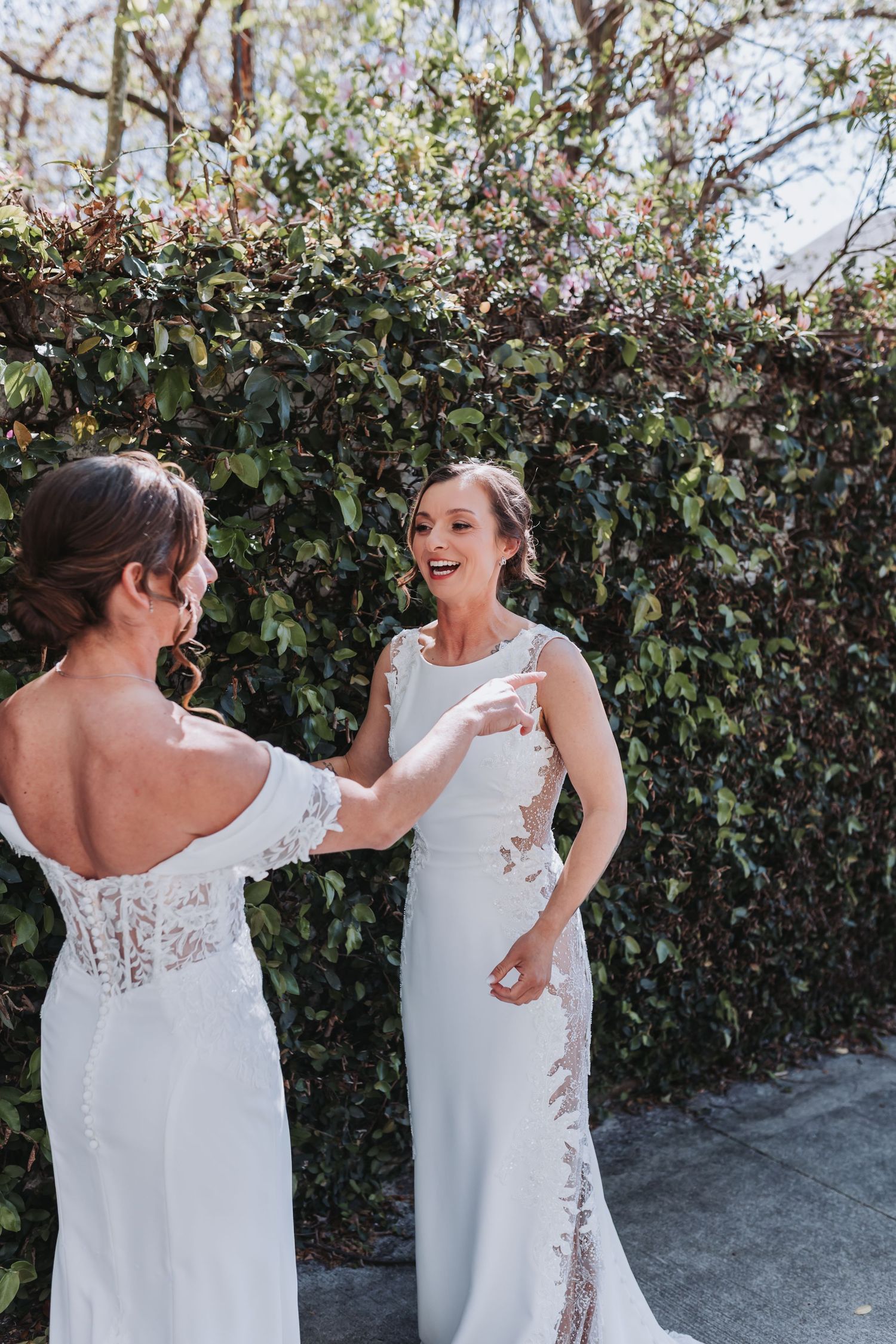 Two brides in elegant lace wedding dresses share a joyful moment outdoors in front of a lush green hedge.