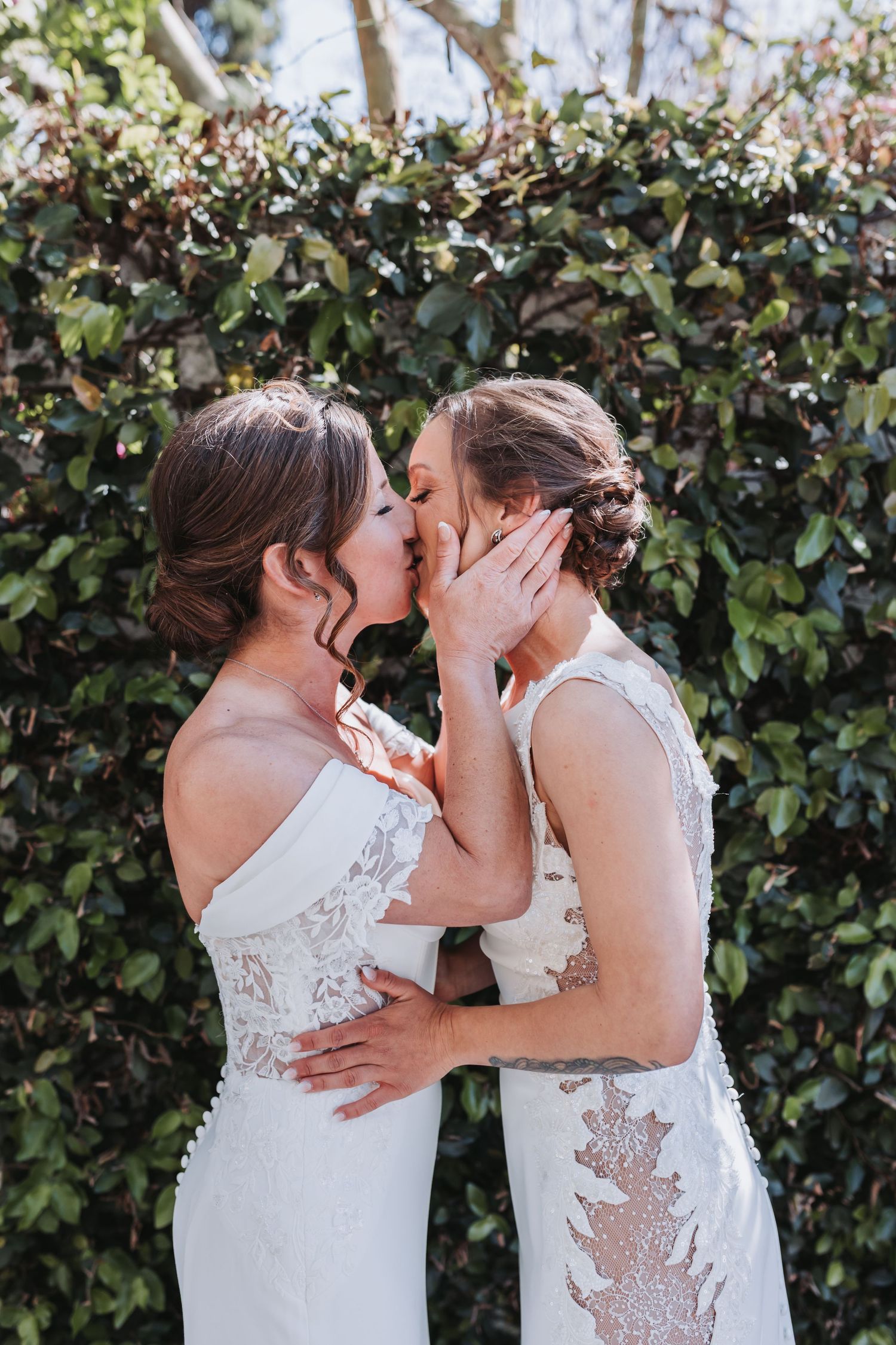 Two brides in white lace wedding gowns share a romantic kiss in front of a lush green hedge.