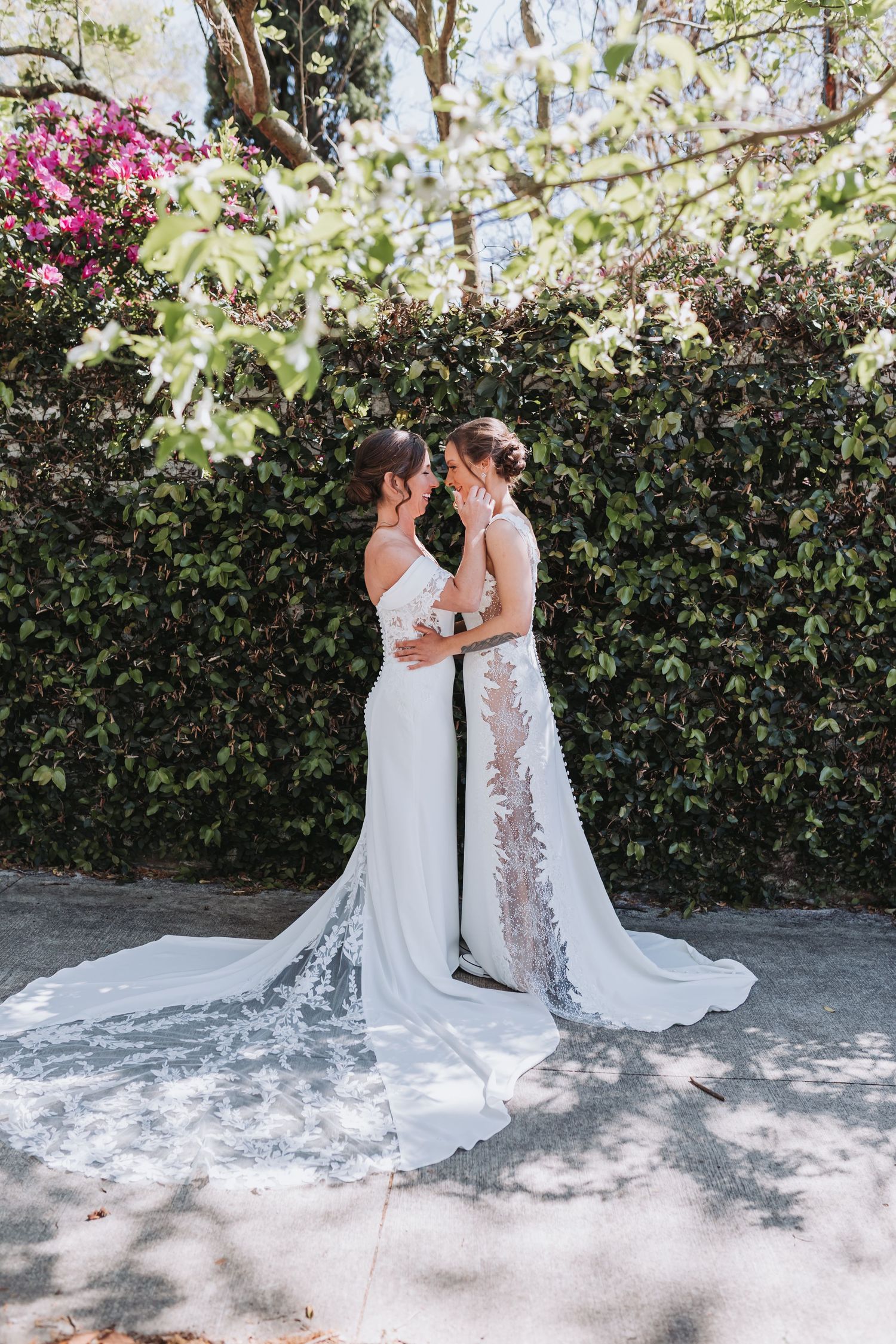 Two brides in white wedding gowns share an intimate moment surrounded by lush green ivy and pink flowers.