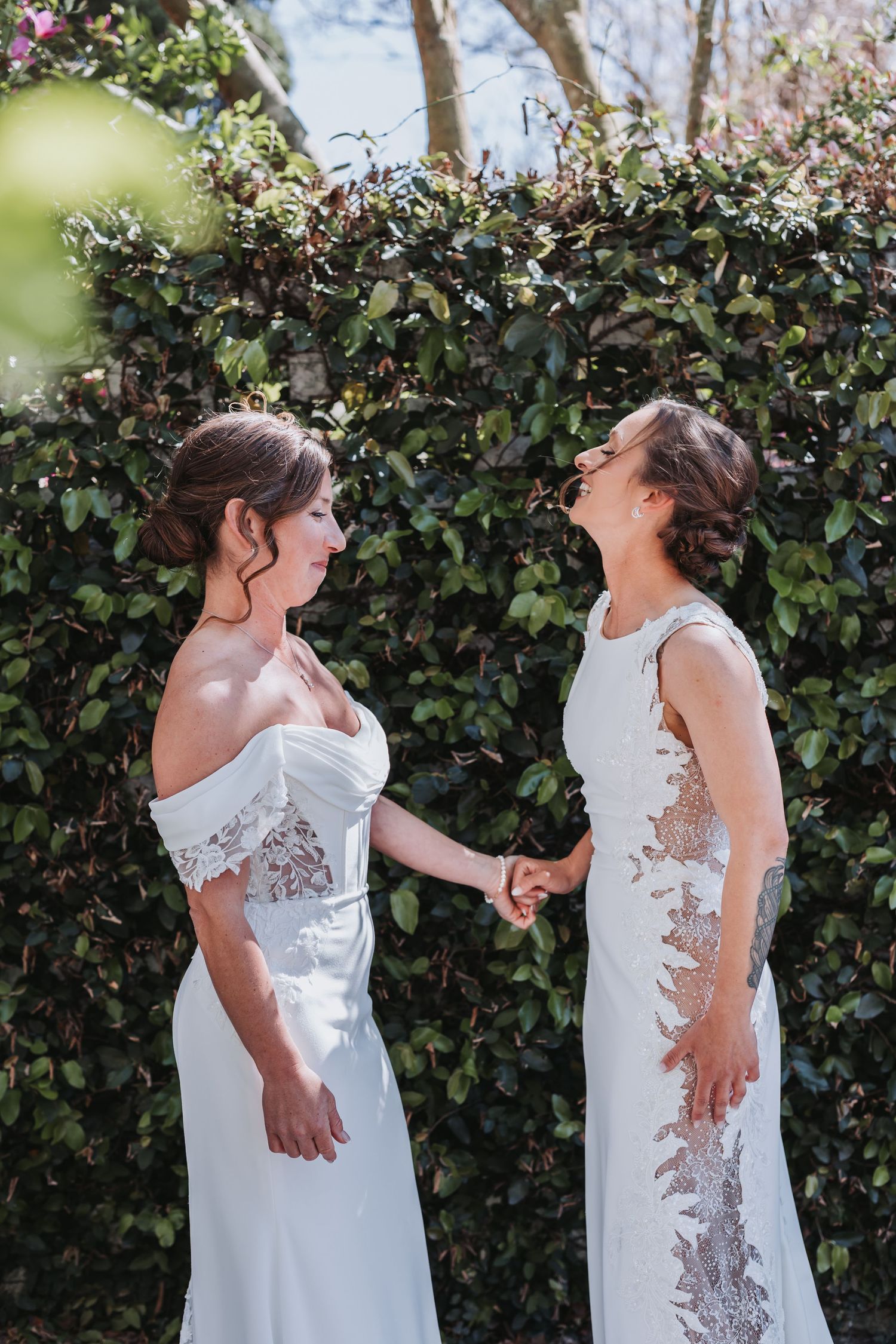 Two brides in white gowns hold hands and smile at each other in front of a lush green hedge backdrop.