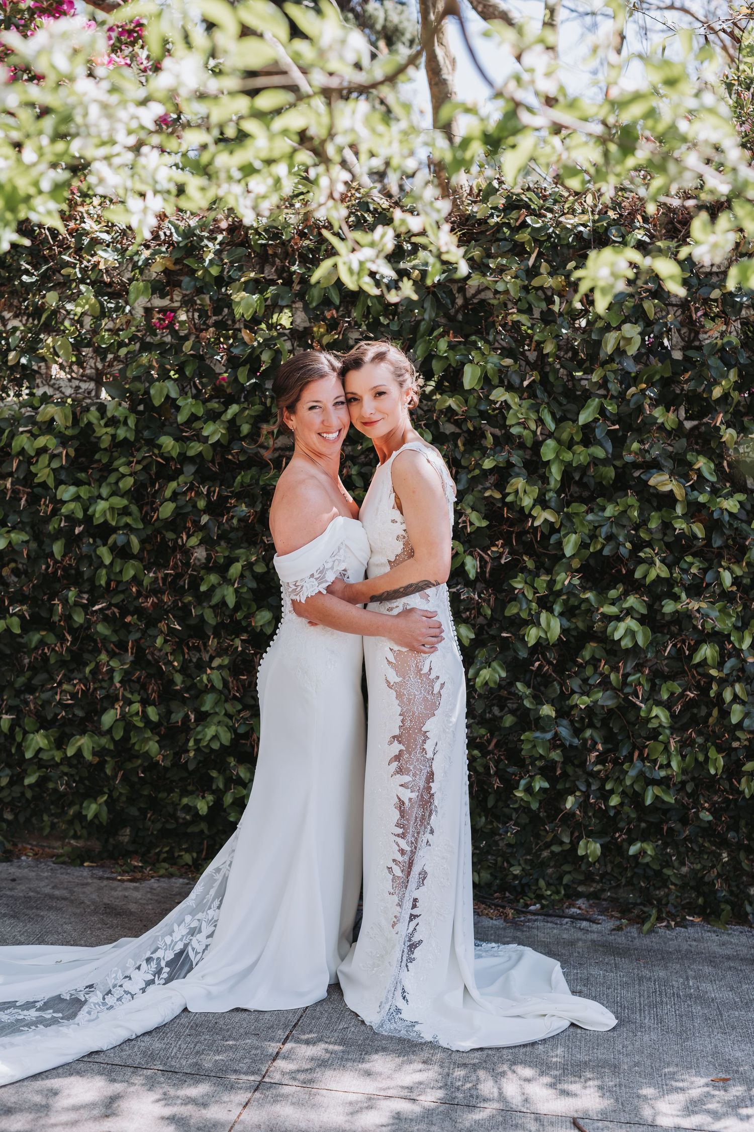 Two brides in elegant white wedding dresses embrace and smile before a blooming green hedge.