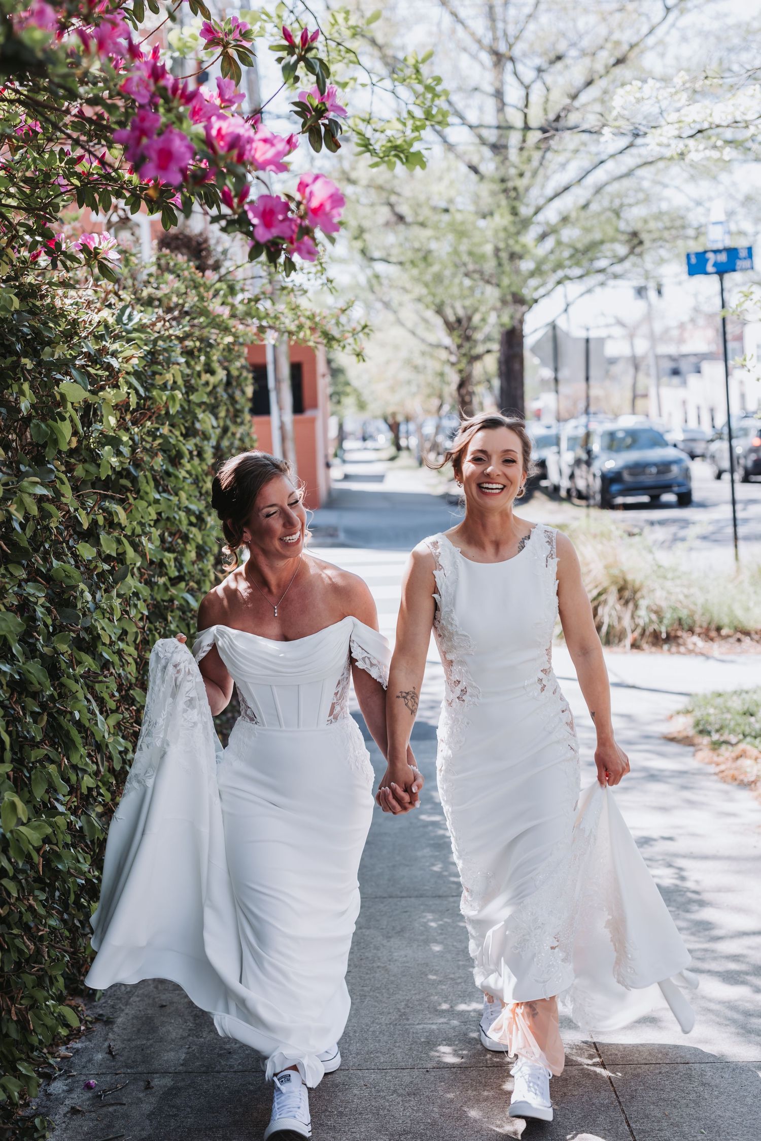 Two brides in white wedding dresses hold hands and laugh while walking past spring blooms.