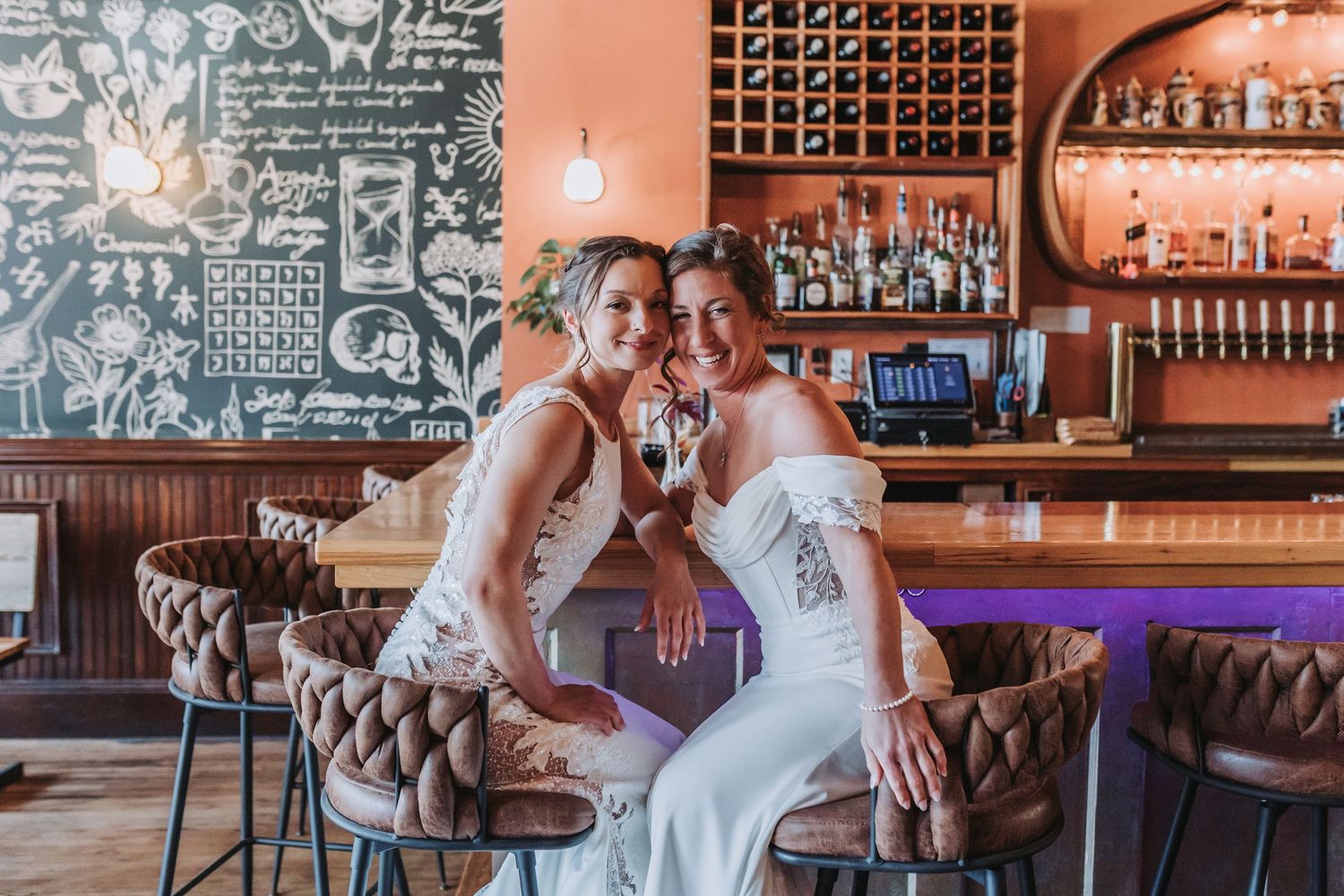 Two brides in white wedding gowns pose together at a stylish bar with warm lighting and a chalkboard wall.