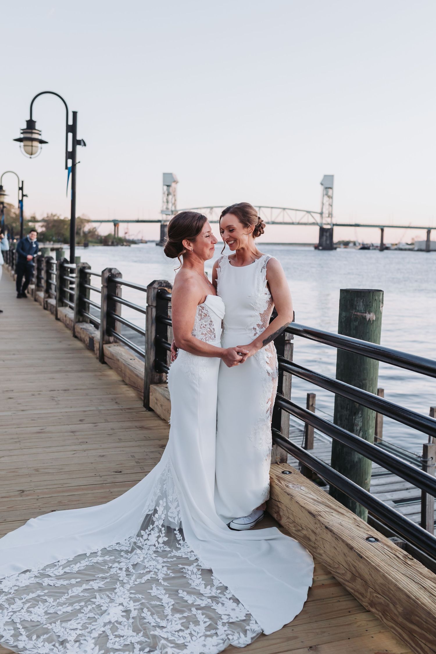 Two brides in elegant white gowns share a joyful moment on a waterfront boardwalk at sunset.