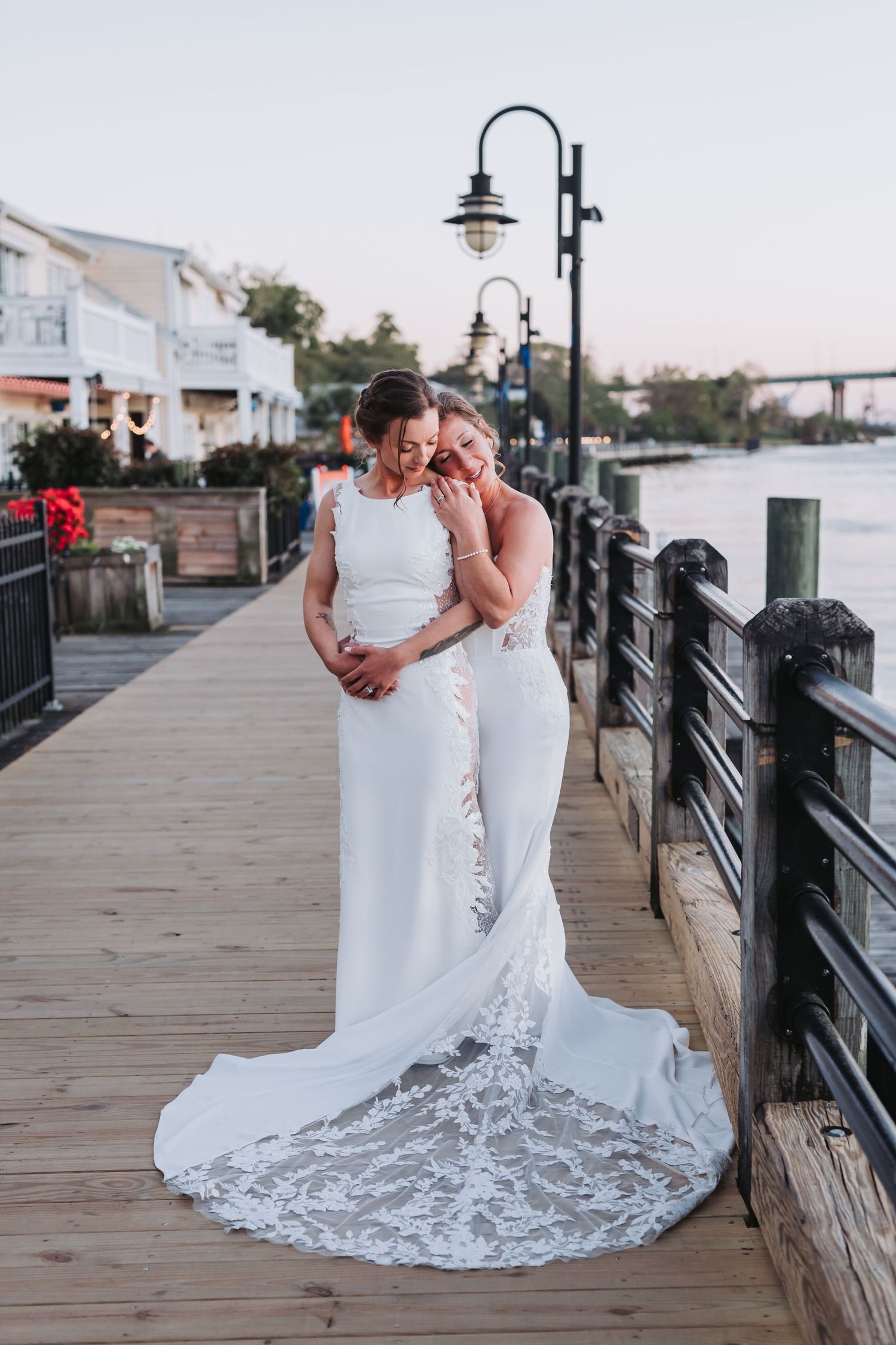 Two brides in white lace gowns share a tender embrace on a waterfront boardwalk at sunset.