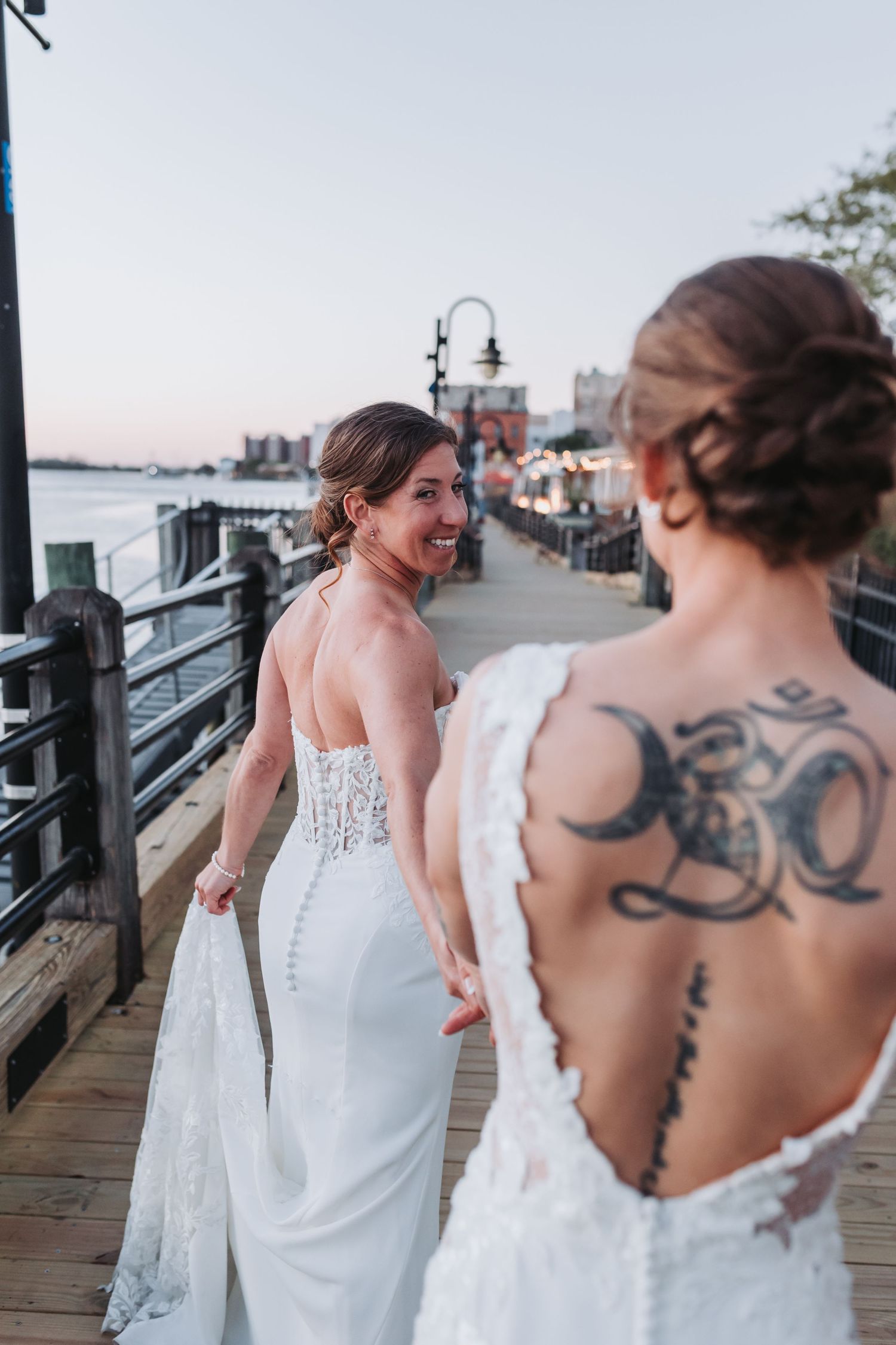 Two brides in white wedding gowns walk hand in hand on a waterfront boardwalk at sunset.