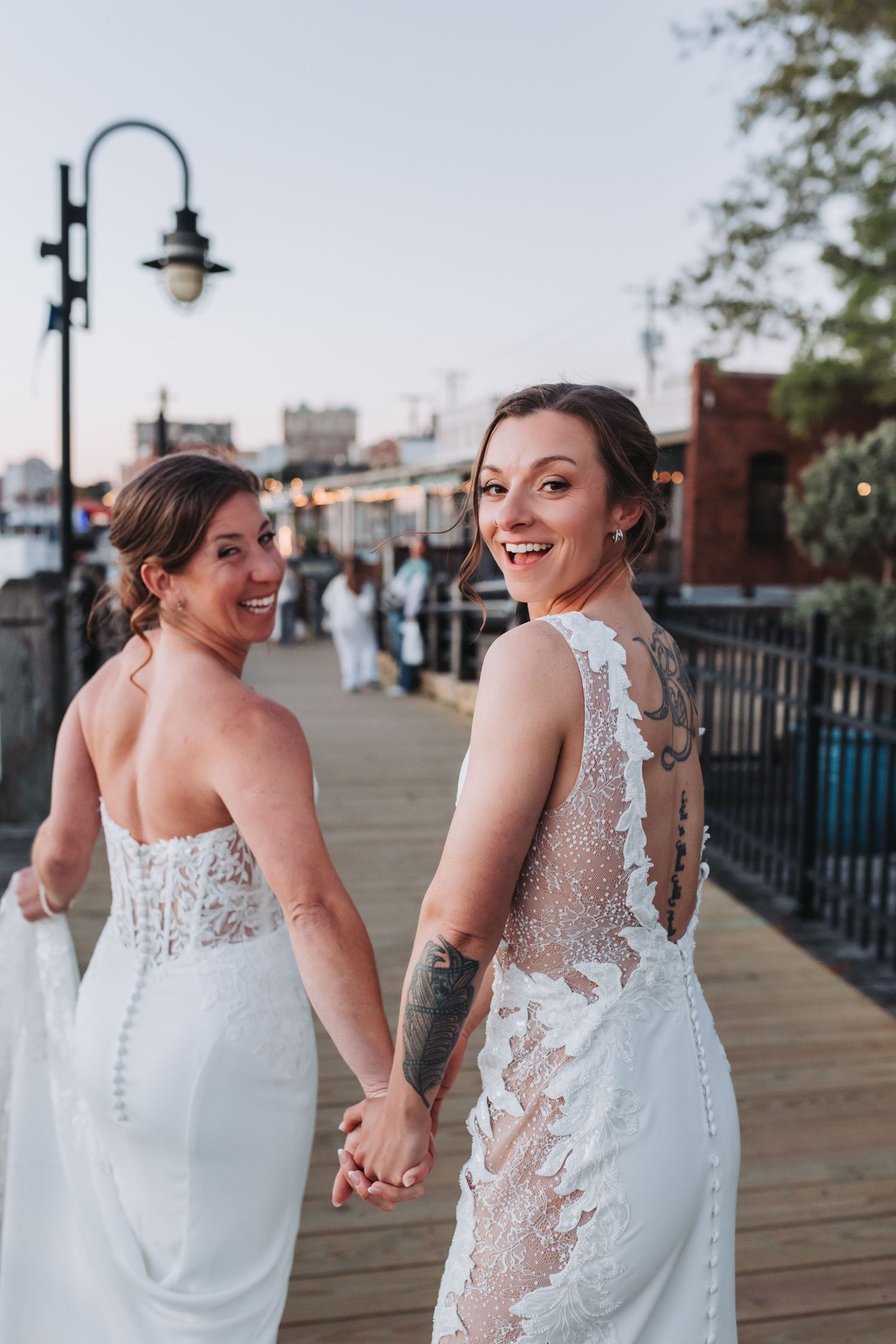 Two smiling brides in white wedding dresses hold hands and look back on a waterfront boardwalk.
