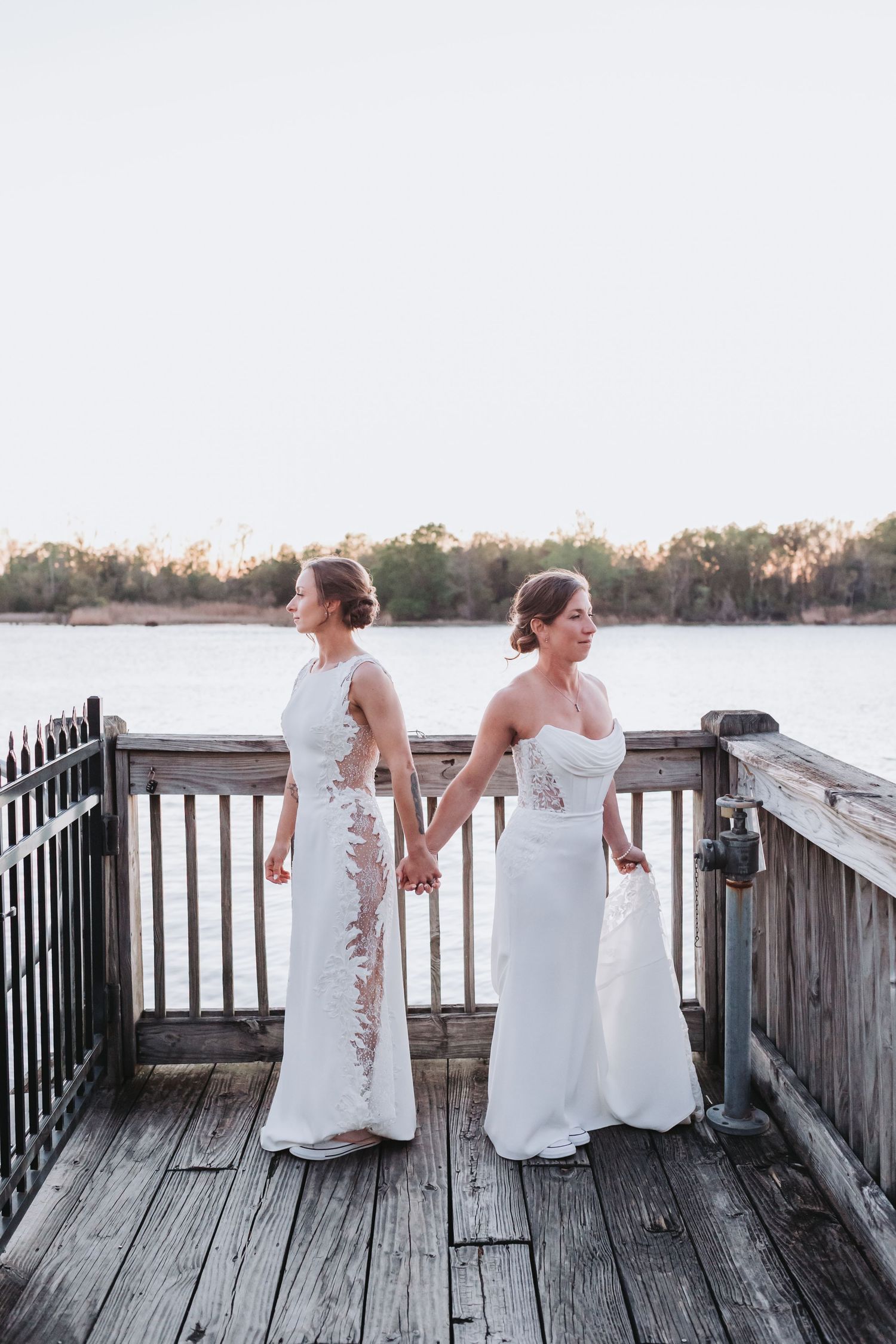 Two brides in white wedding gowns stand hand in hand on a wooden dock overlooking a serene river.