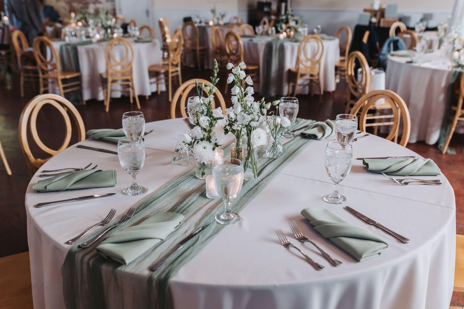 Elegant wedding reception table with sage green napkins, white floral centerpiece, and glassware.