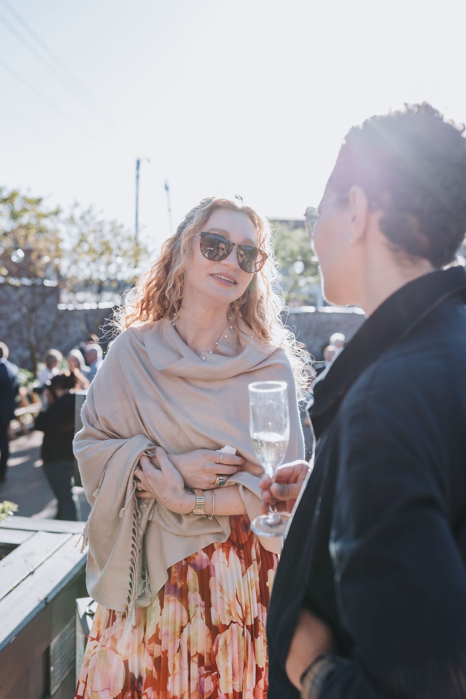 Woman in sunglasses holding champagne flute at outdoor wedding reception on sunny day.