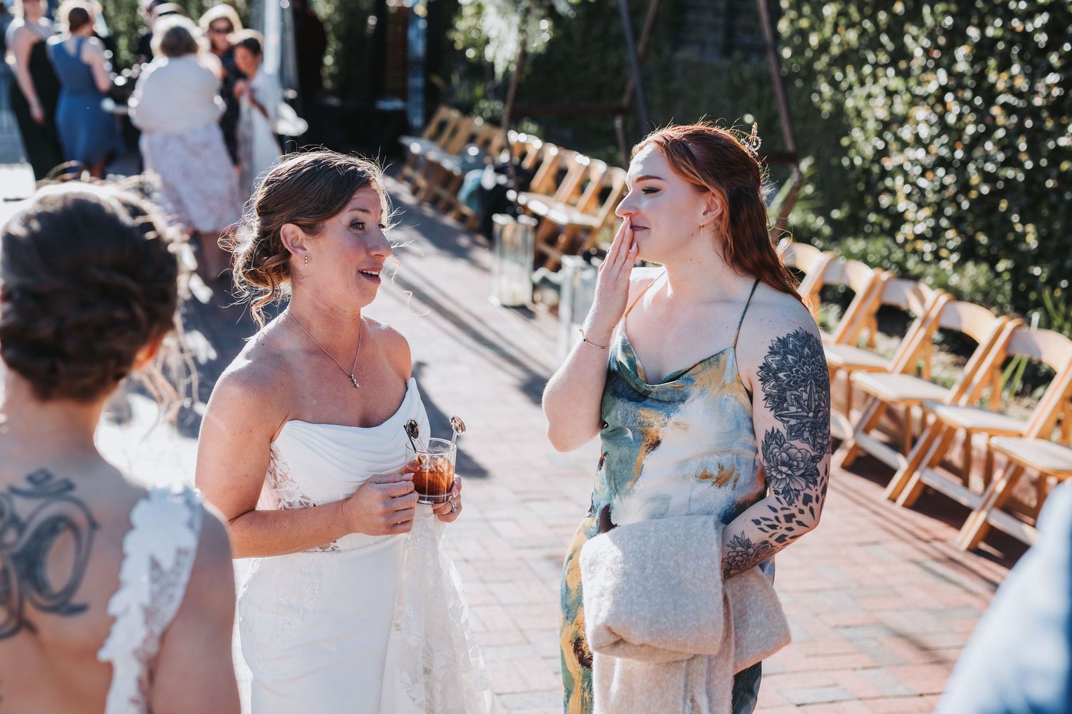 Bride in white gown laughing with tattooed guest at outdoor wedding ceremony.