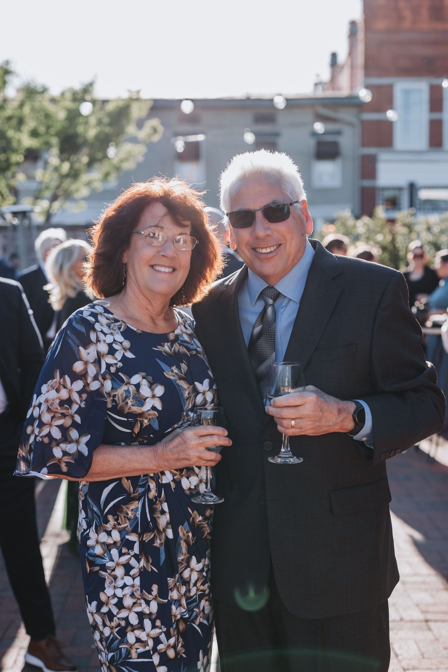Smiling senior couple holding champagne glasses at an outdoor wedding reception on a sunny day.