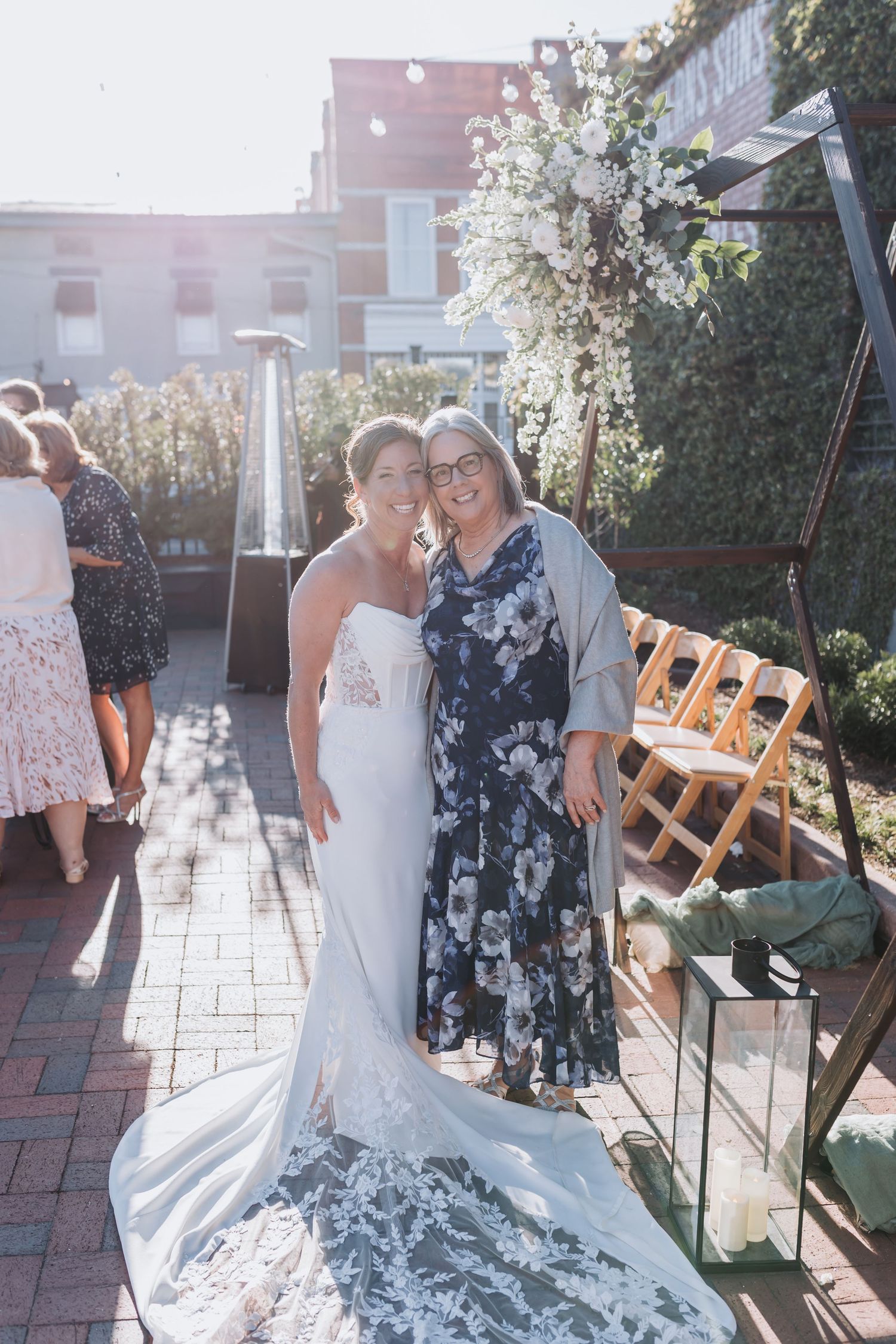 Bride in white lace gown poses with guest in floral dress at sunny outdoor wedding venue.