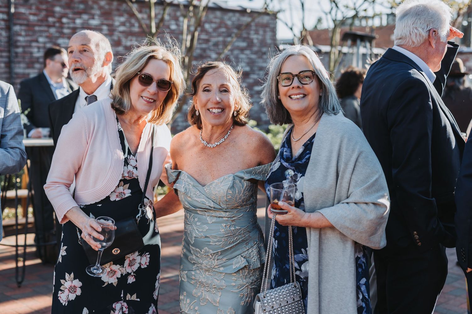 Three women smiling and holding drinks together at an outdoor wedding reception celebration.