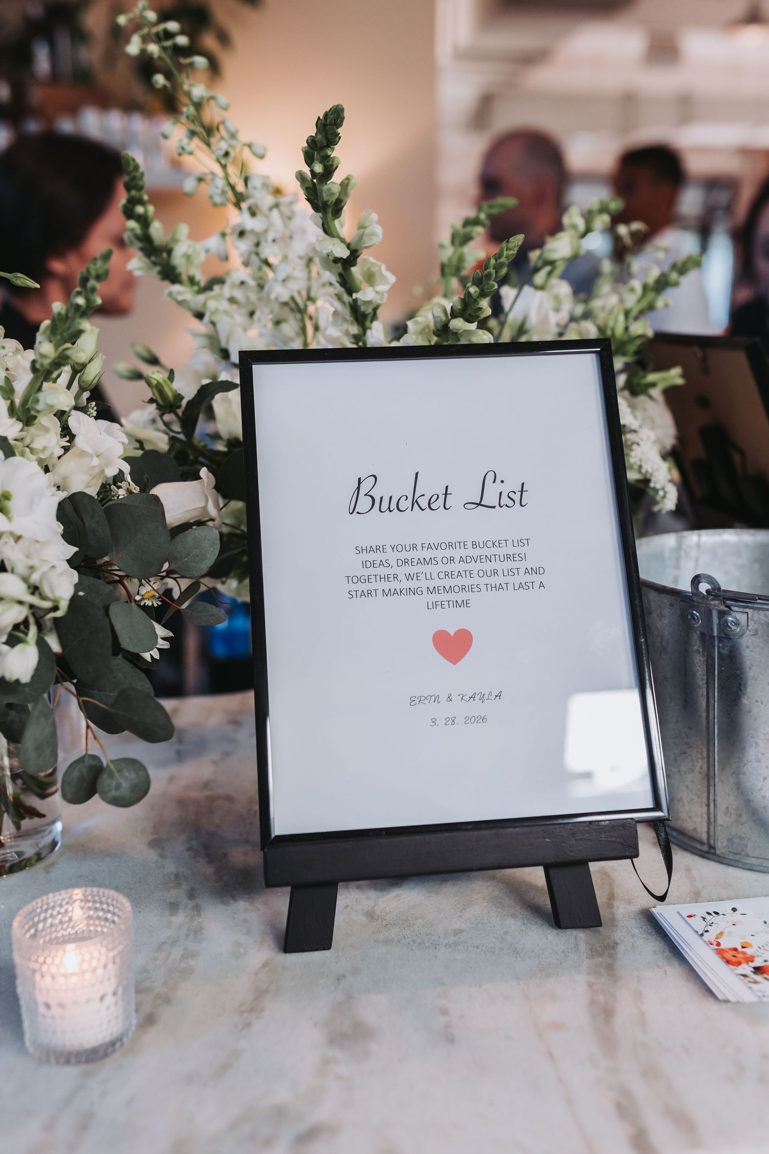 Framed wedding bucket list sign displayed on easel with white flowers and candles at reception table.