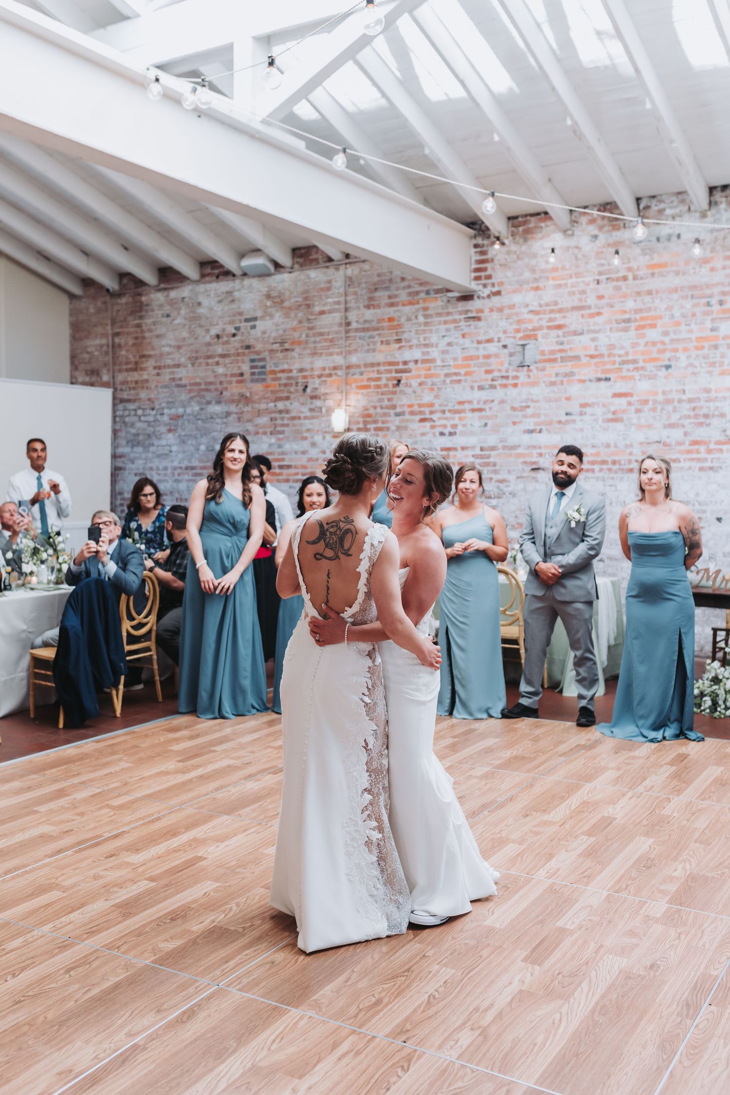 Two brides share first dance in white gowns on wooden floor surrounded by wedding party in dusty blue dresses.