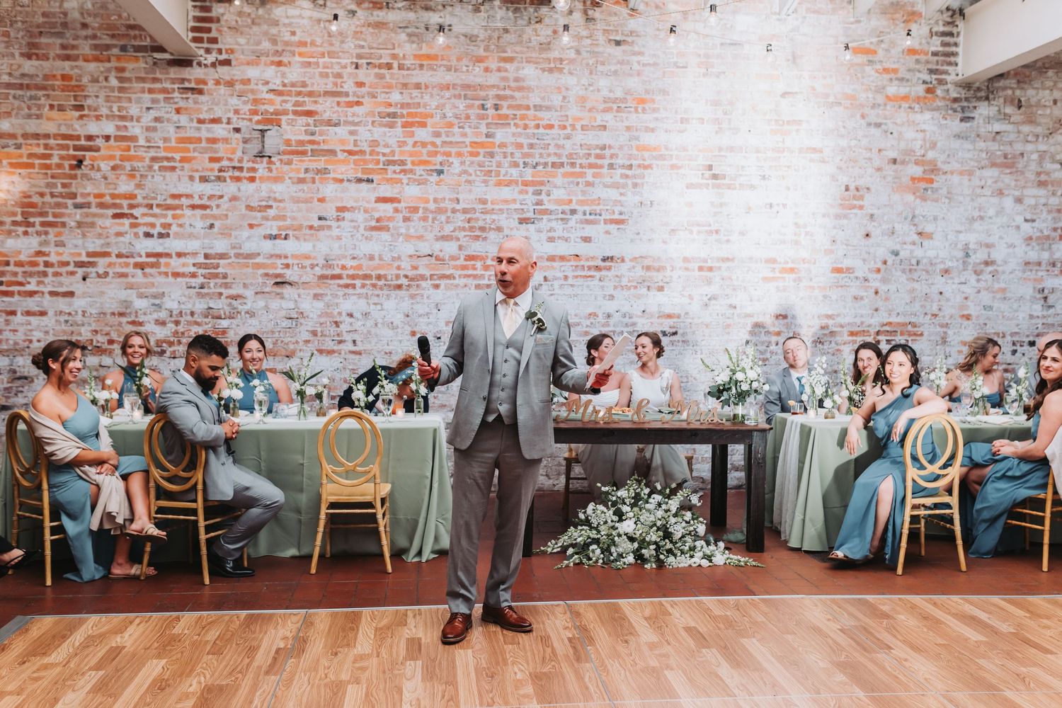 Bald man in gray suit giving a wedding toast speech to seated guests at a reception.