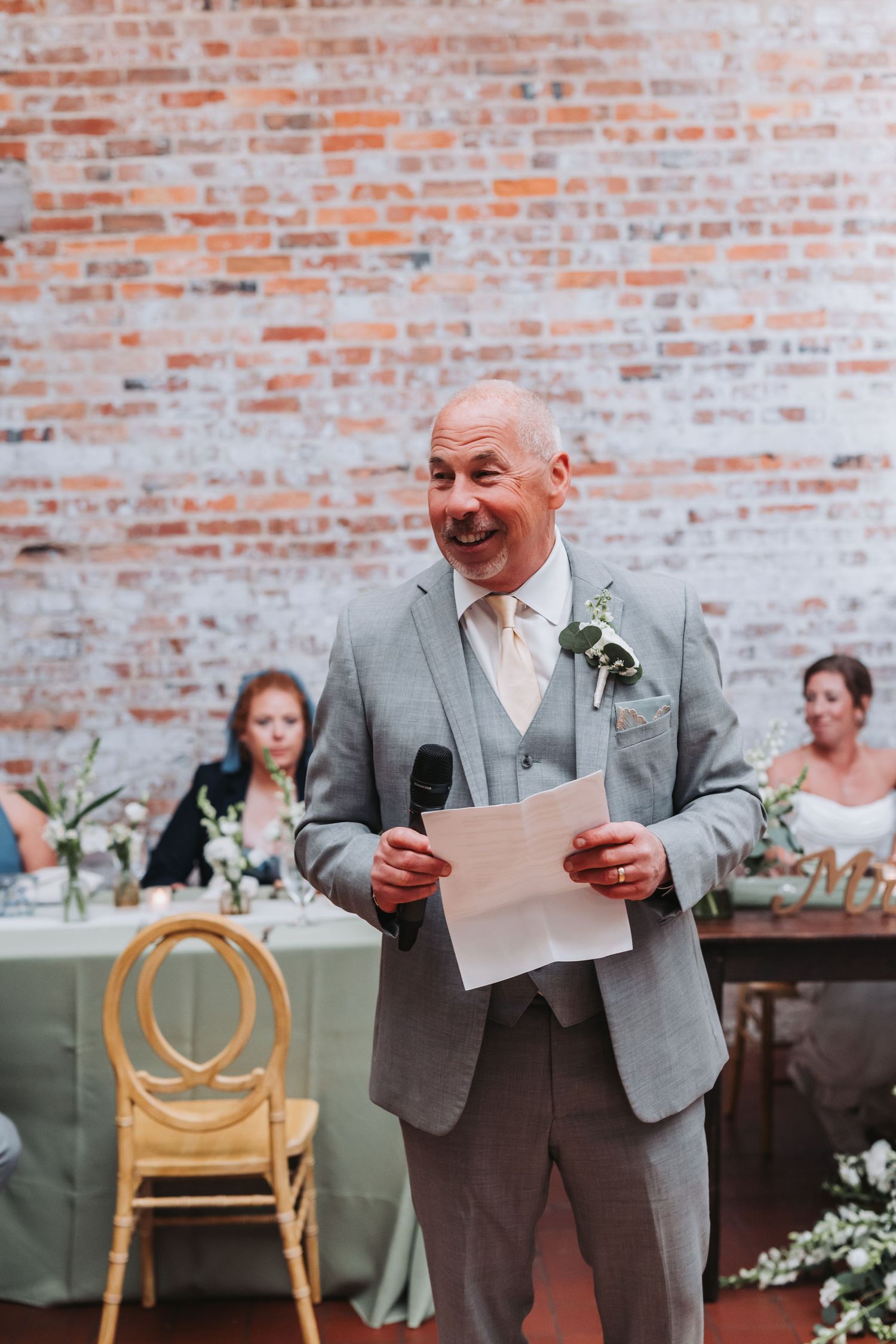 Smiling bald man in gray suit giving wedding speech holding microphone and paper in front of brick wall.