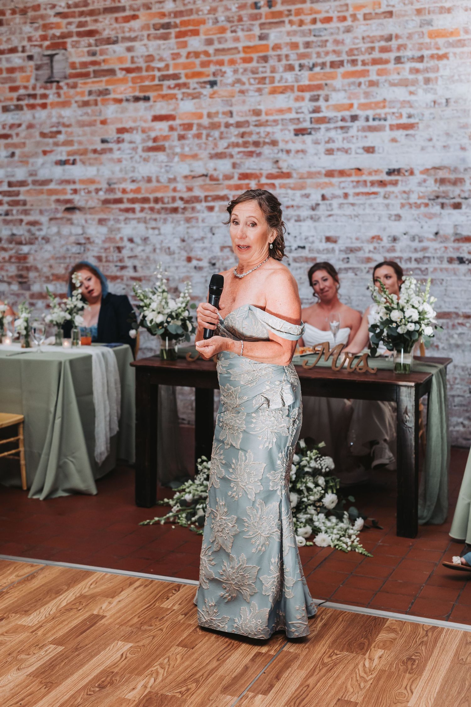 Elegant woman in embroidered blue gown giving wedding speech with microphone in front of exposed brick wall.