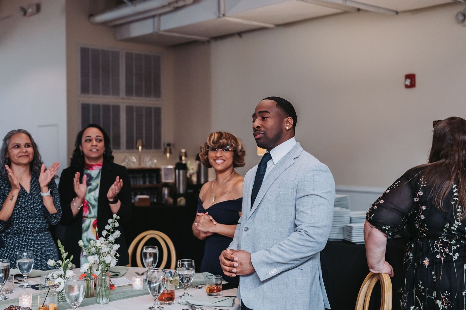 A man in a light gray suit stands and claps with guests during a wedding reception in an elegant venue.