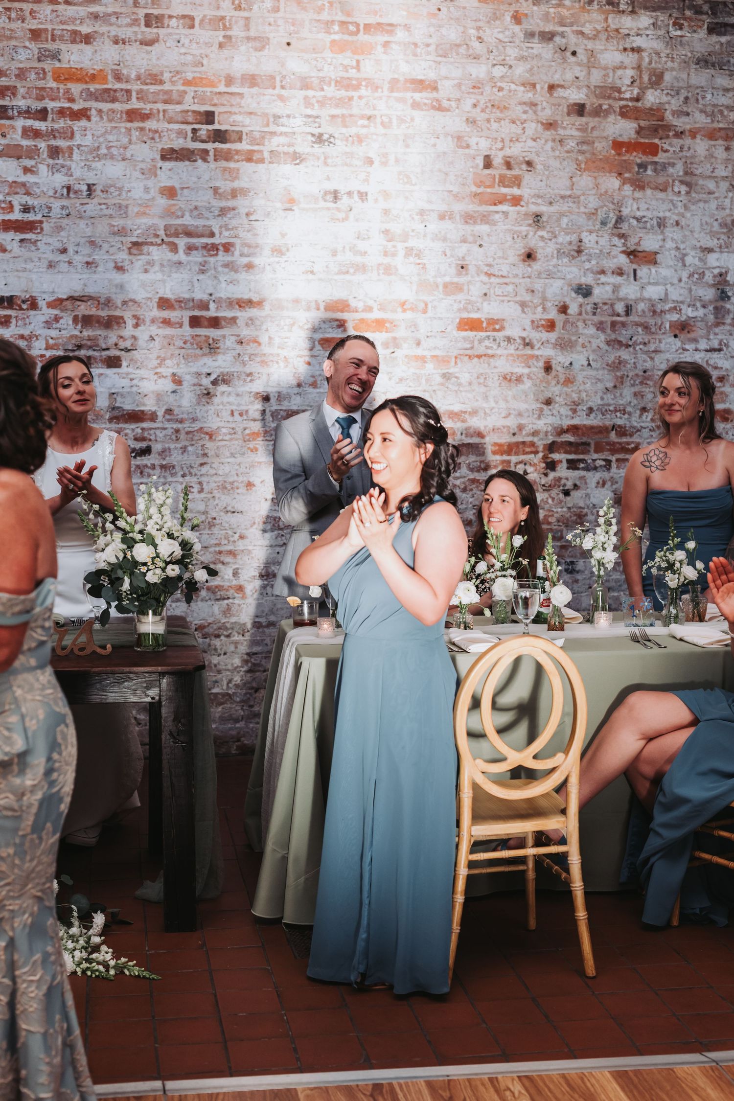 Bridesmaid in blue gown clapping at wedding reception table against exposed brick wall backdrop.