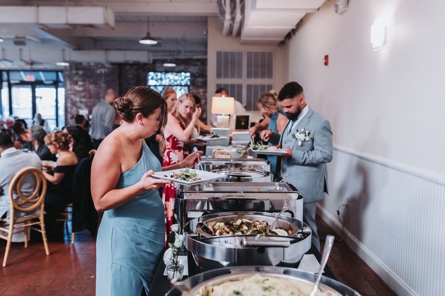 Wedding guests serving themselves at a buffet line with chafing dishes in a brick-walled reception venue.