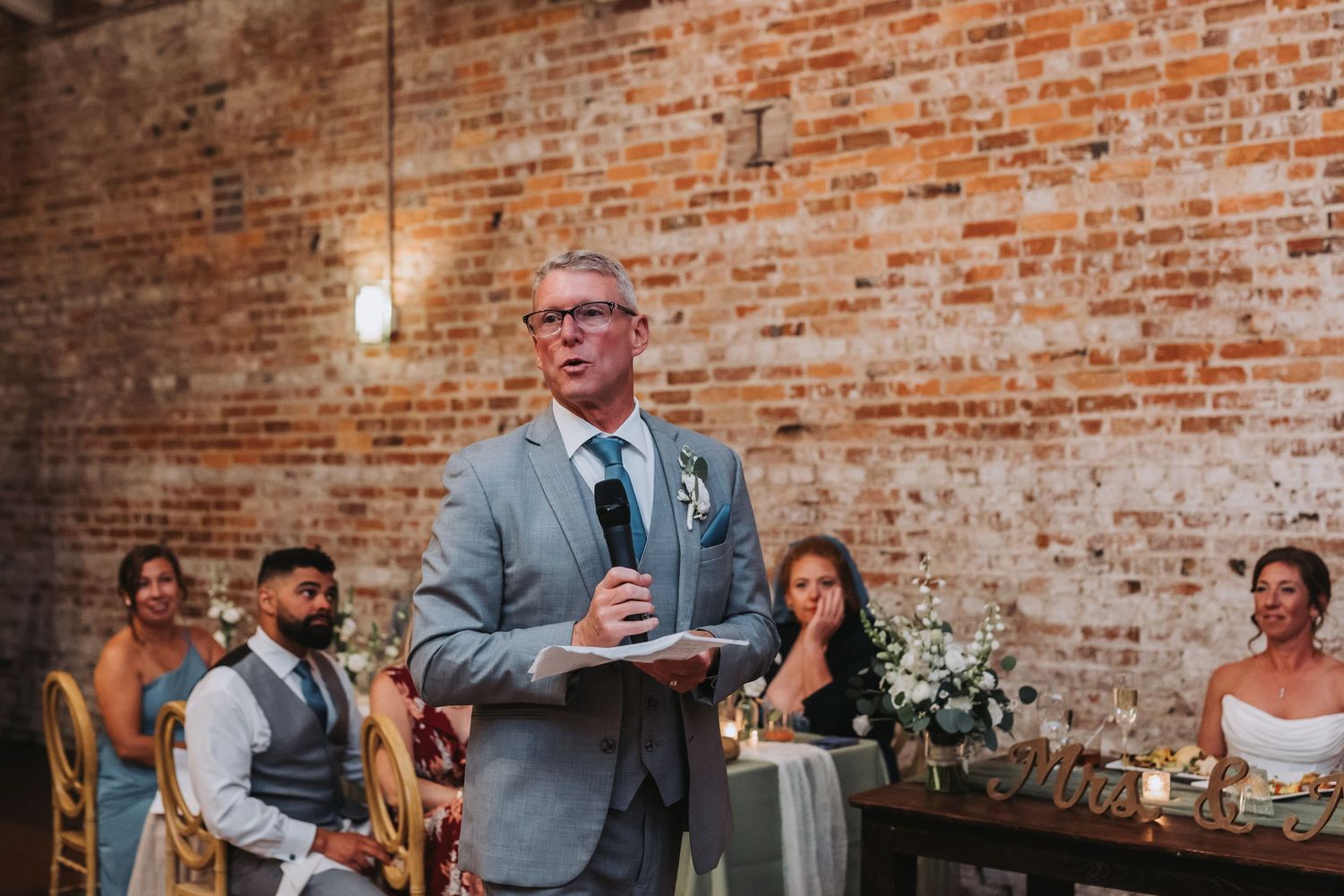 Man in gray suit delivering wedding speech at microphone in front of exposed brick wall at reception.
