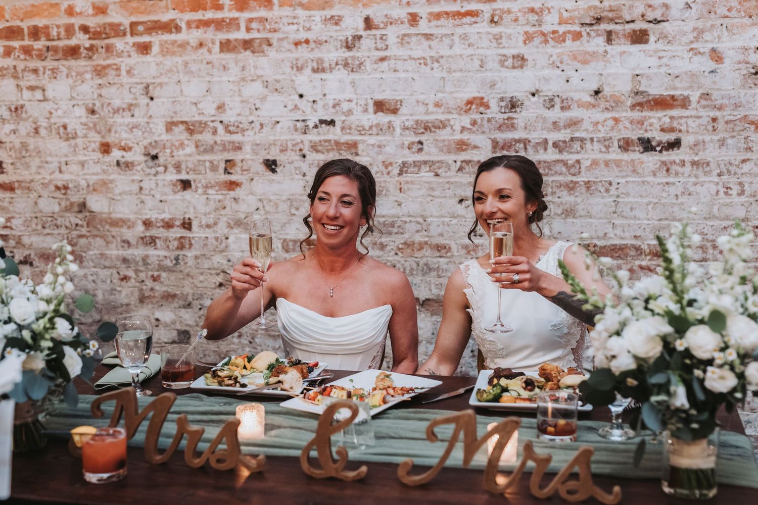 Two brides toasting champagne at Mrs and Mrs sweetheart table with white florals.
