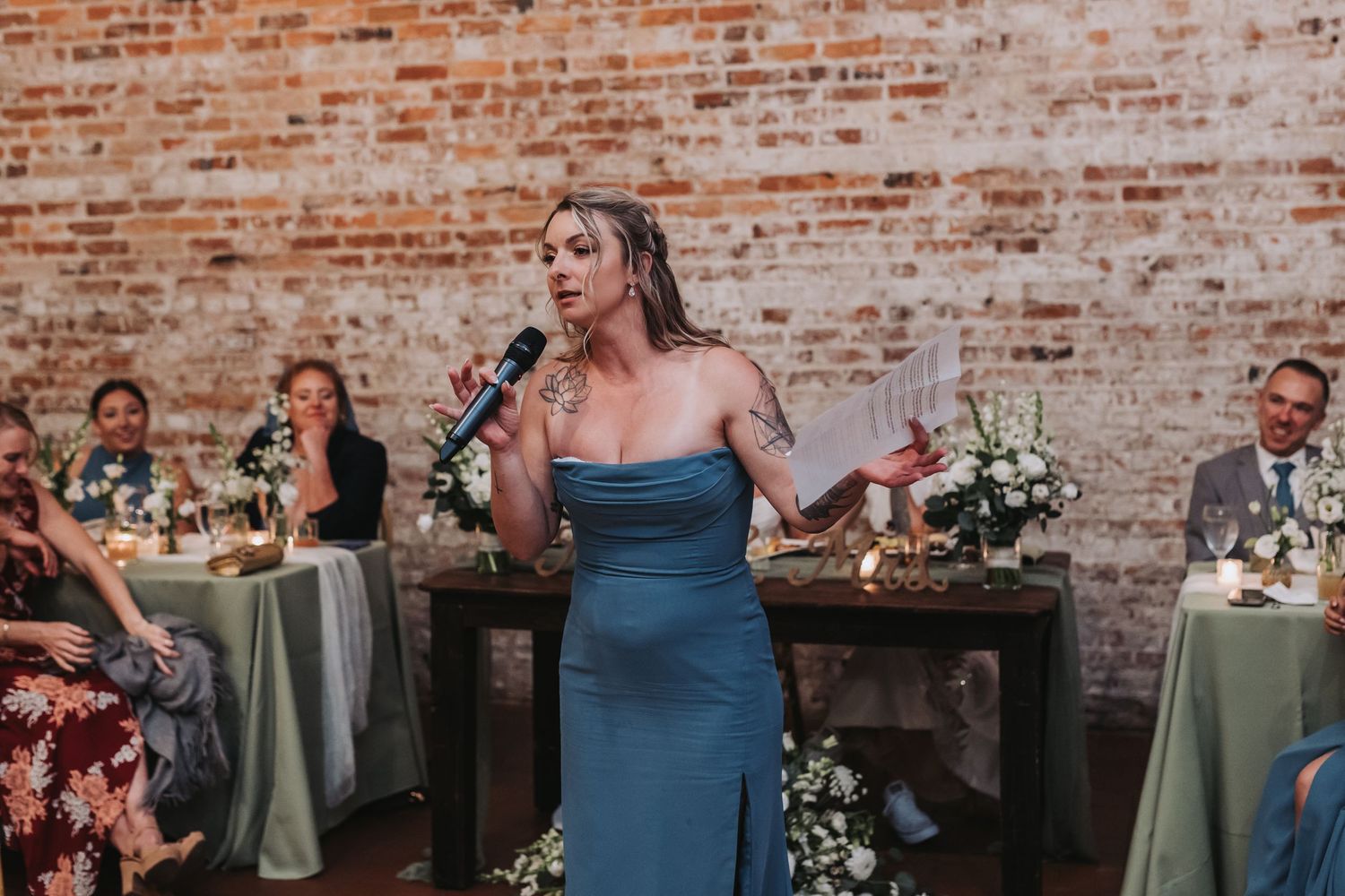 Woman in blue strapless gown giving an emotional wedding toast speech at brick-walled reception venue.