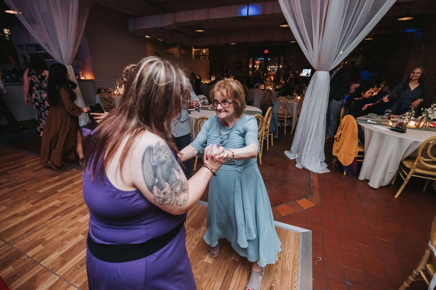 Two women dance together at wedding reception in draped venue with wooden dance floor.